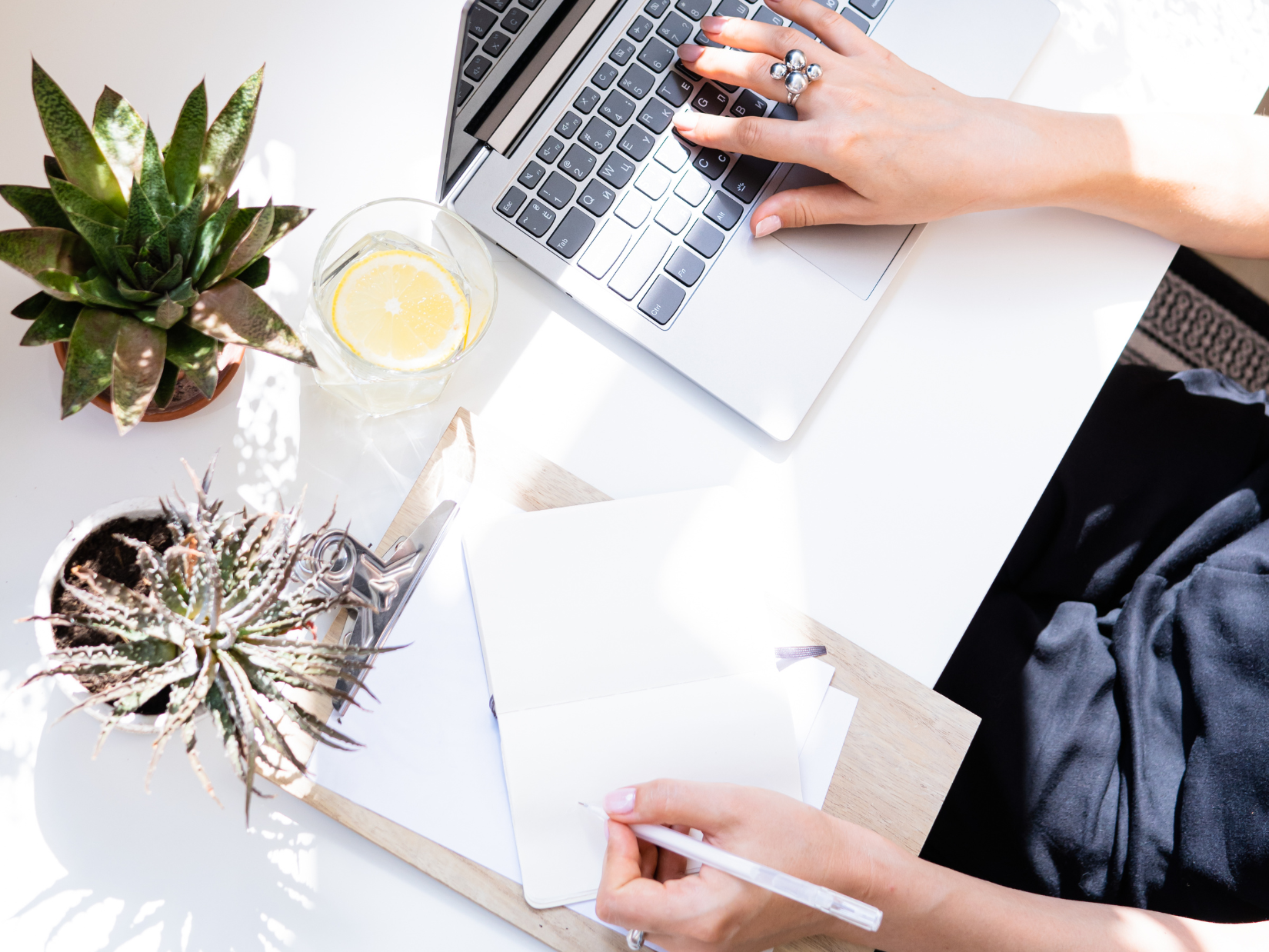 A person working on a laptop at a white desk with two potted succulents, a glass of water with lemon, a notepad, and a pen.