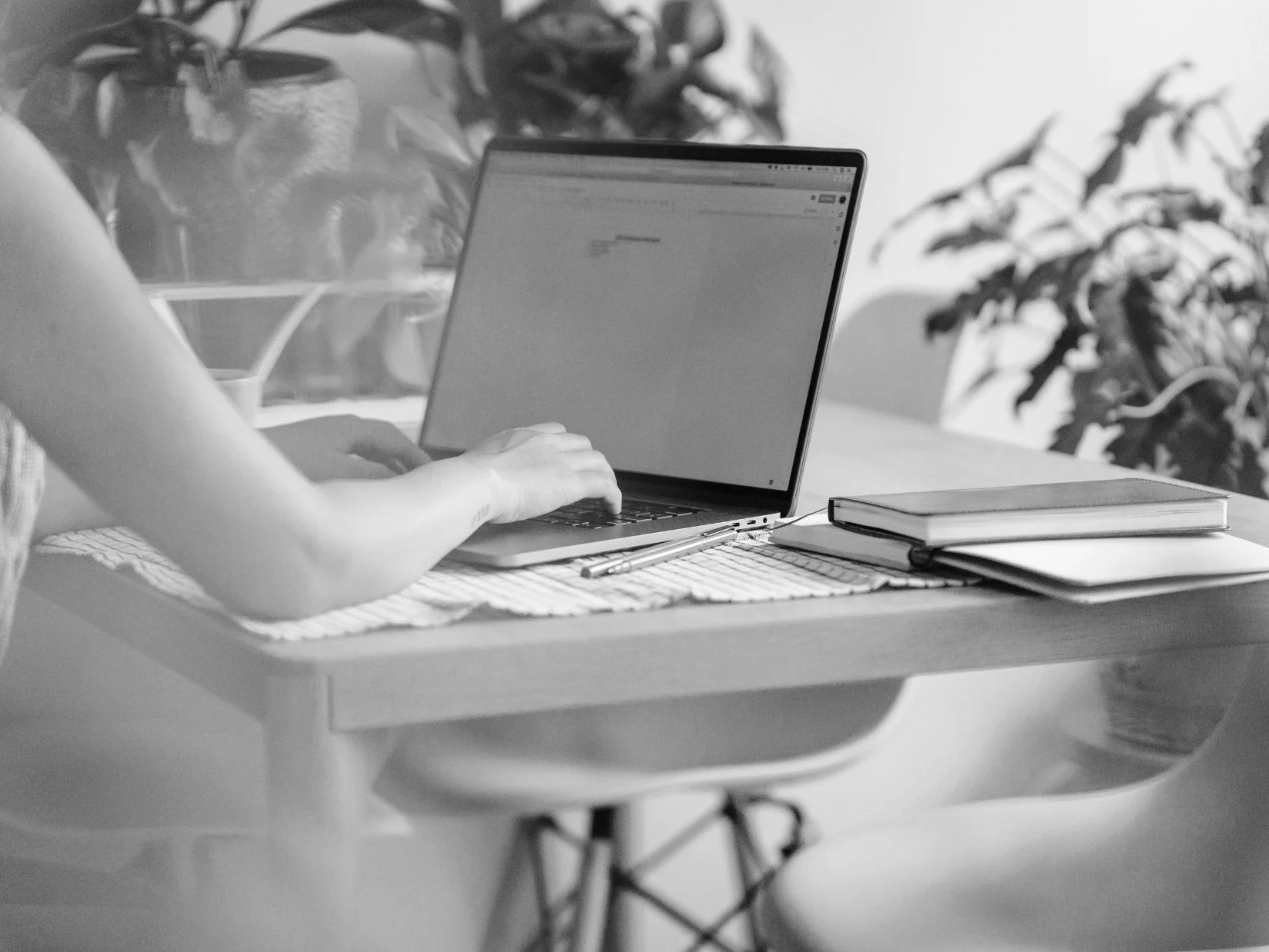 A person working on a laptop at a desk with notebooks and a pen, in a room with plants in the background.