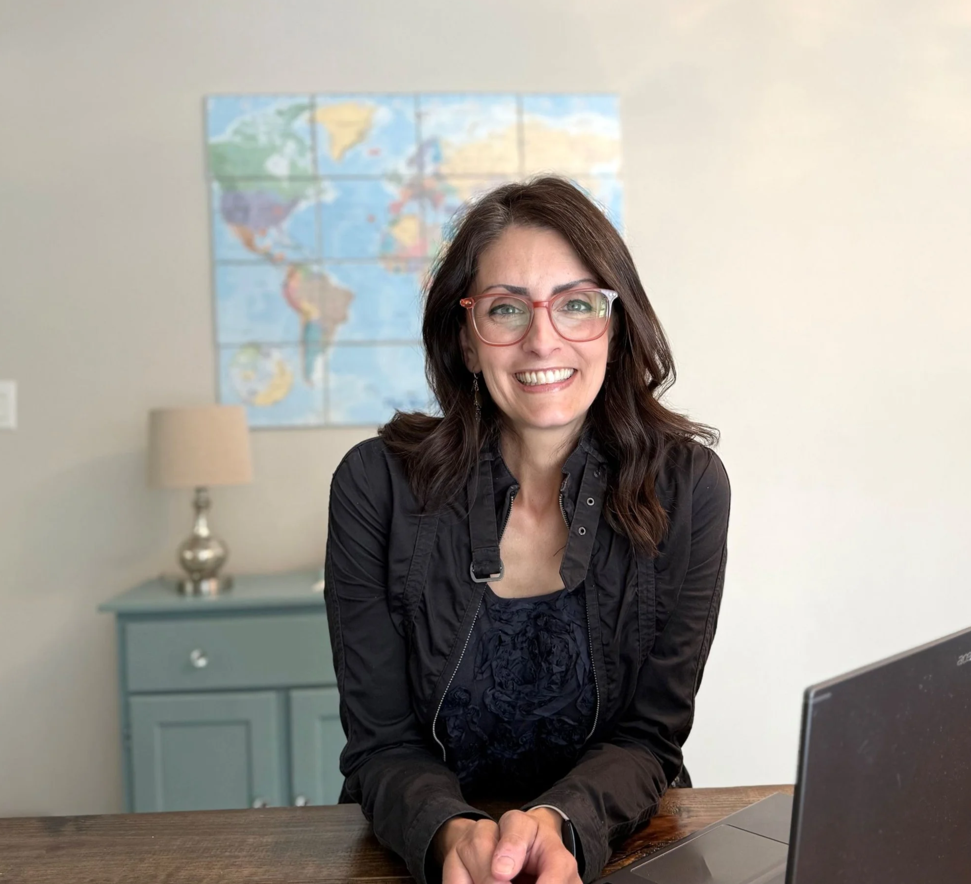 elizabeth daniels in dark grey jacket and pink glass smiling at wooden table with open laptop to her side