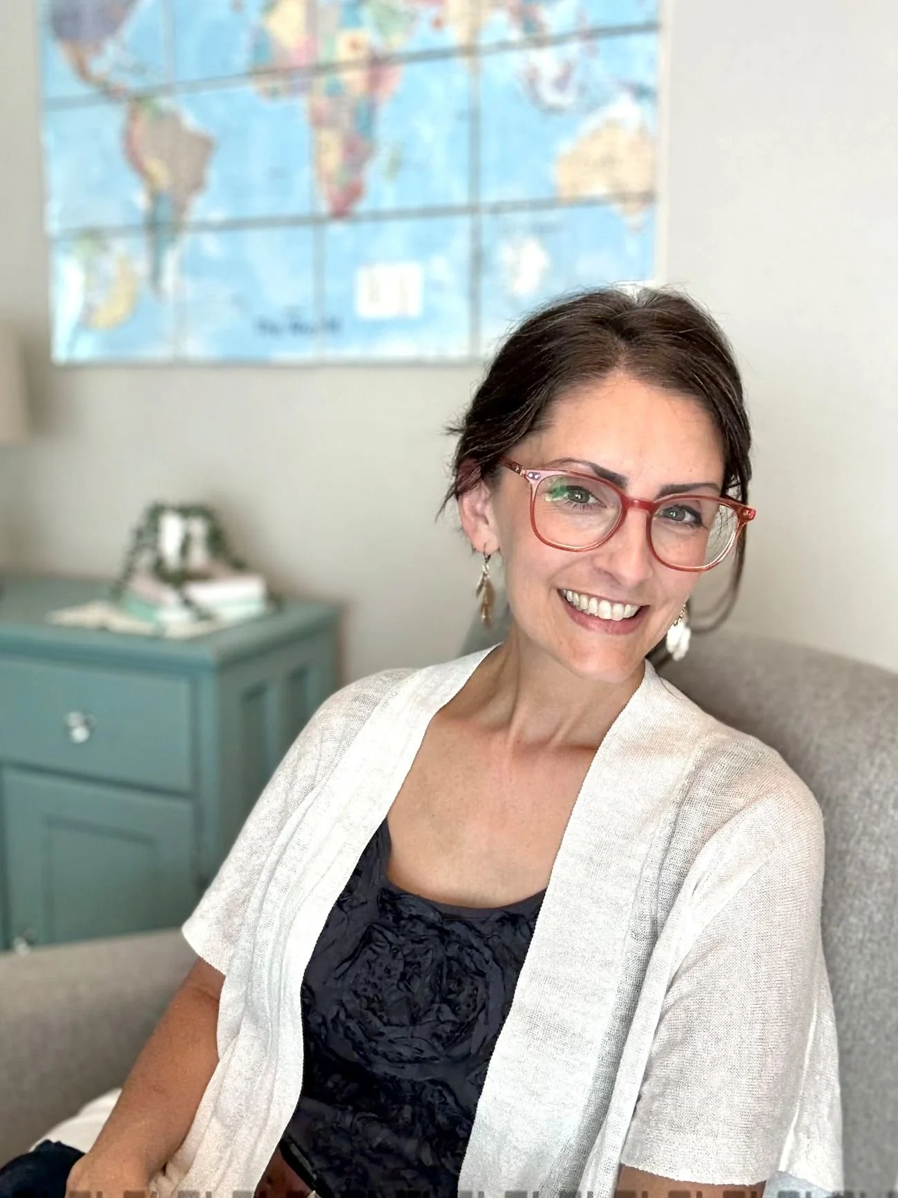 Elizabeth Daniels sitting on a beige armchair in a room with a blue side table, a stack of books, and a world map on the wall in the background, smiling.
