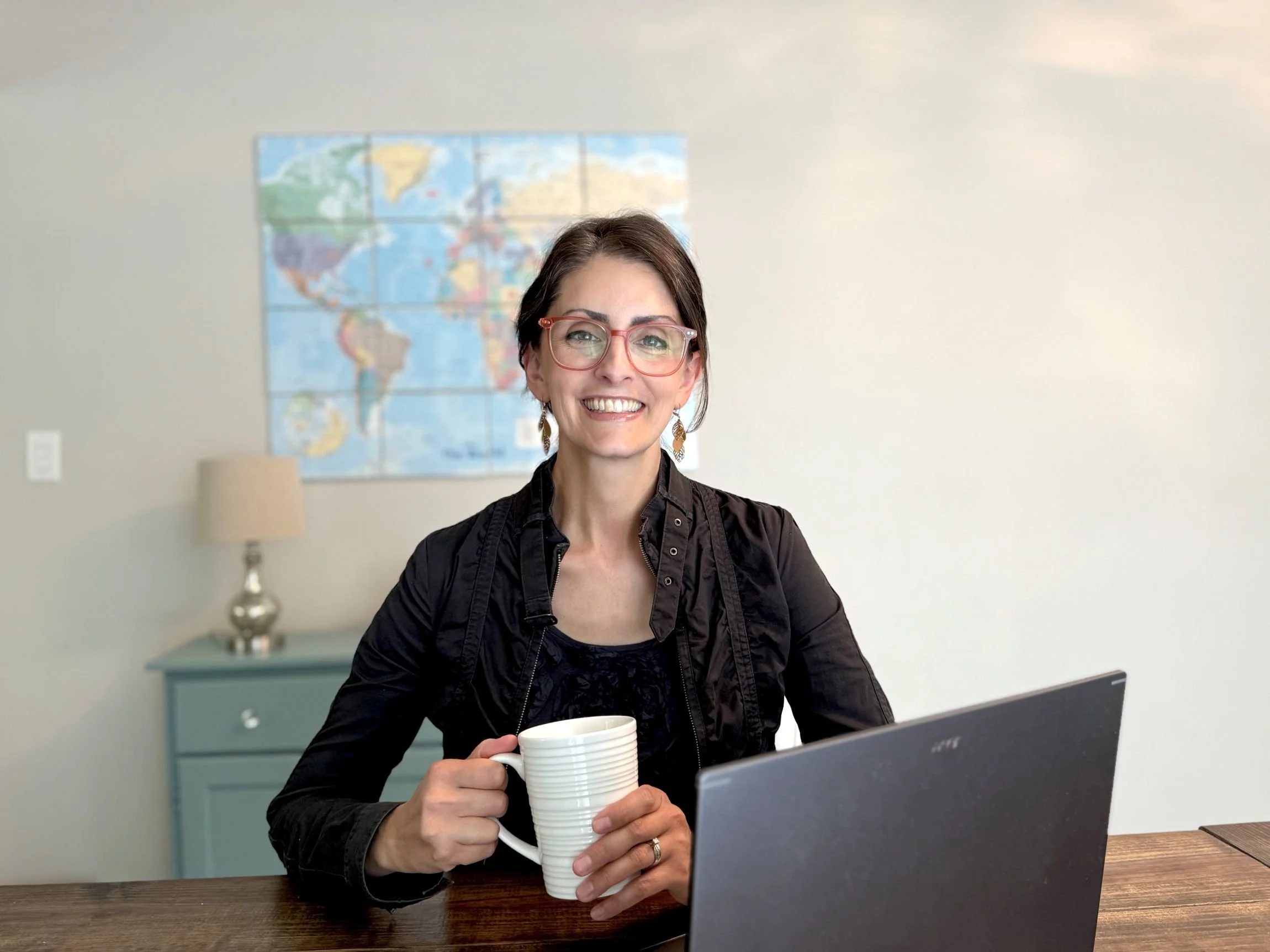 elizabeth daniels sitting at open laptop smiling with white mug in hand and world map behind her