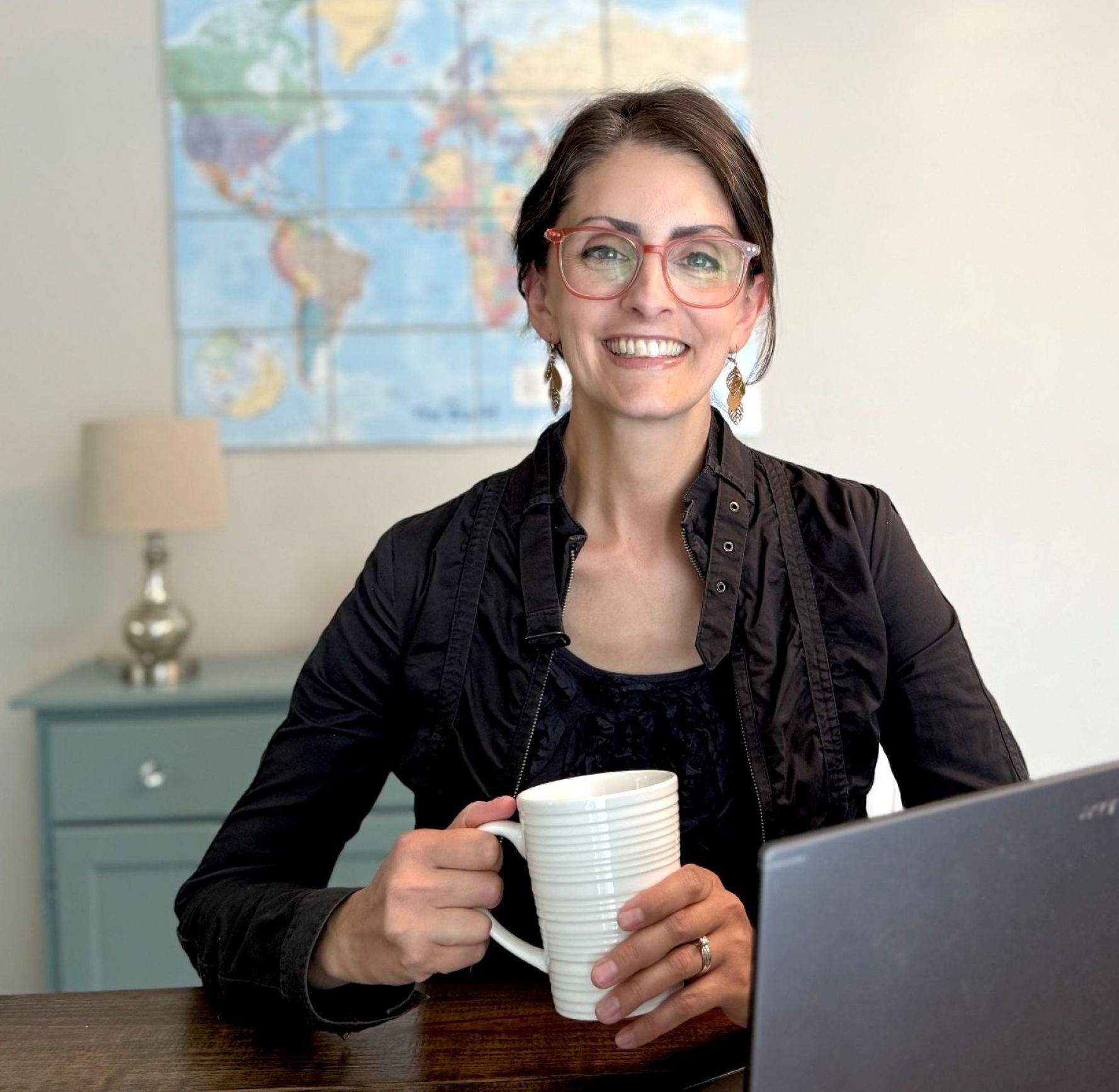 elizabeth daniels in pink glasses holding white mug sitting behind laptop smiling