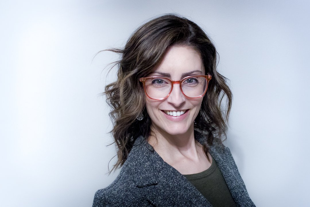 headshot of elizabeth daniels in pink glasses with grey blazer wavey short brown hair
