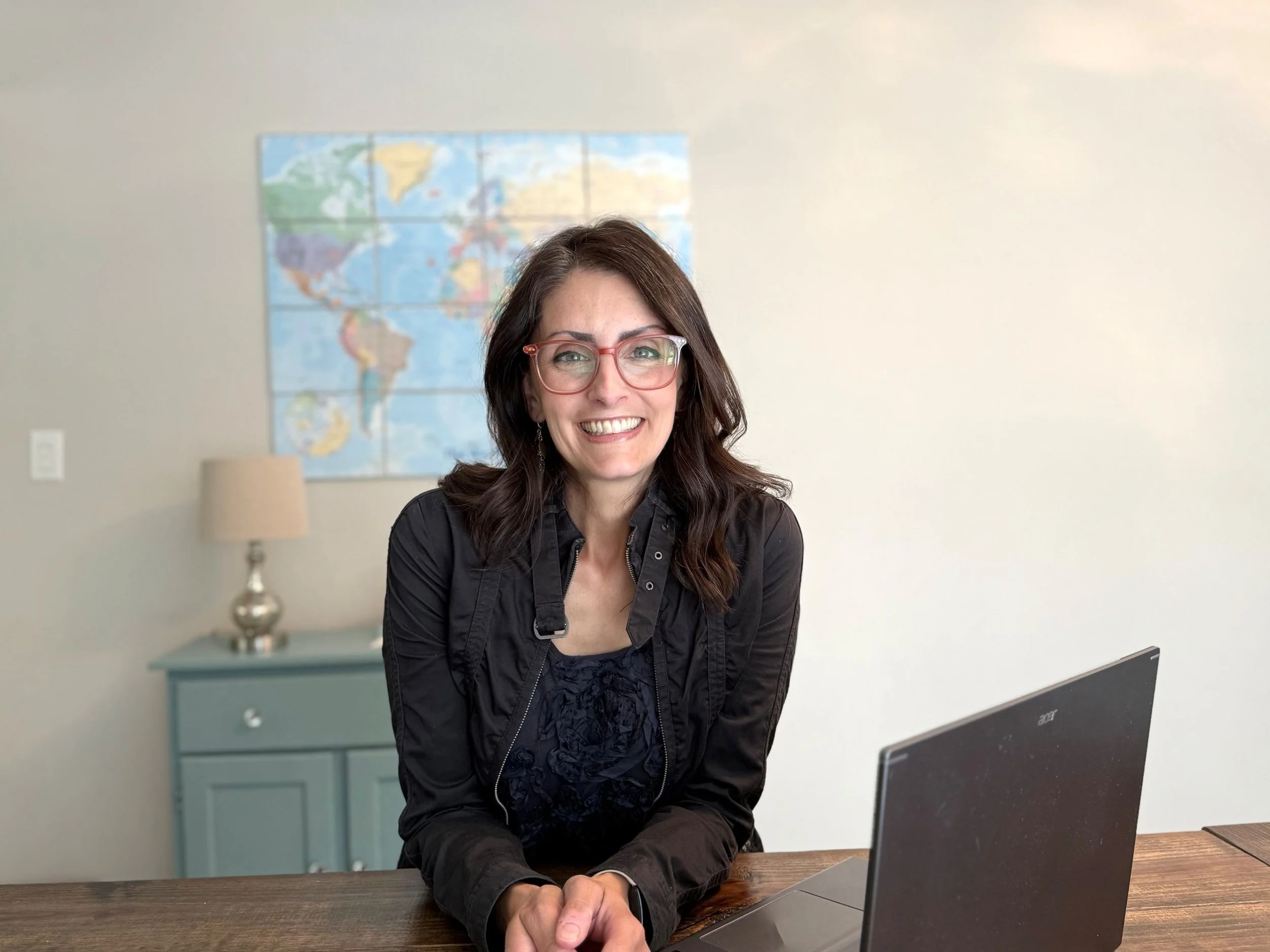 elizabeth daniels leaning on wooden table next to open laptop smiling world map on wall behind