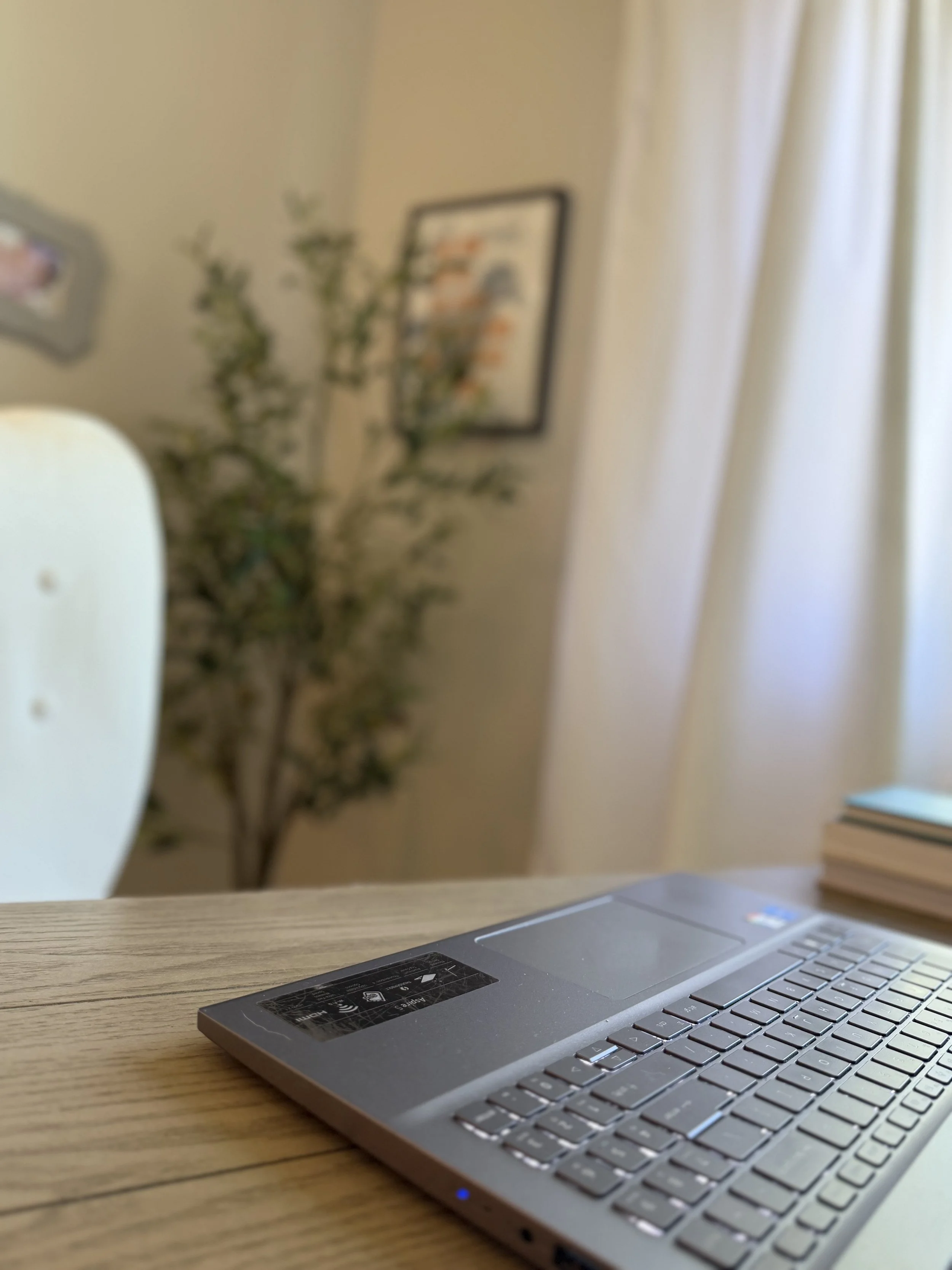 Close-up of a silver laptop keyboard on a wooden desk with a blurred background of a wall, framed picture, curtain, and a stack of books.