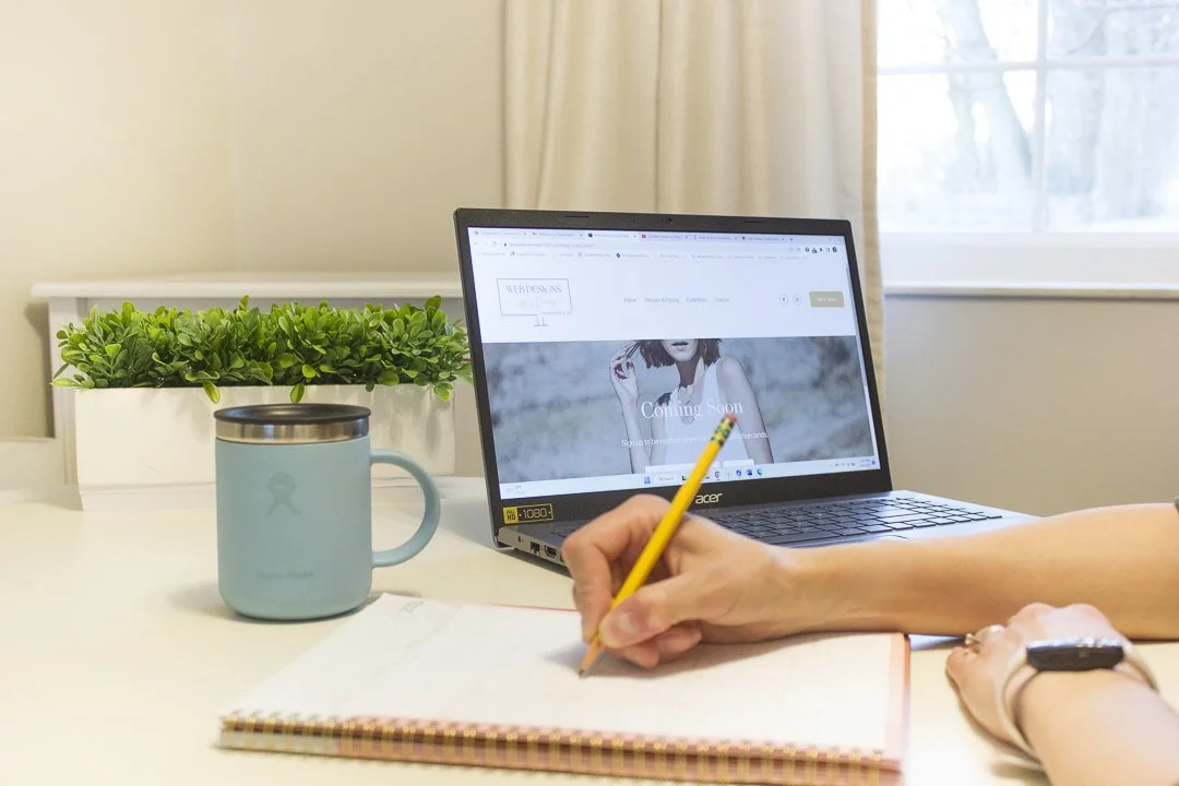 A laptop on a desk displaying a webpage with a woman and the text 'Coming Soon', a person's hand writing in a notebook with a yellow pencil, a light blue mug, and a green plant in the background near a window.