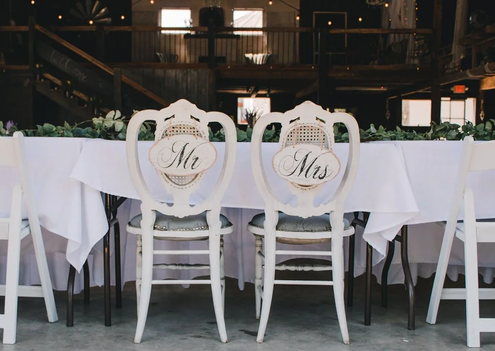 Wedding reception table with two white chairs labeled 'Mr' and 'Mrs' in elegant script, set in rustic venue with wooden beams and string lights.