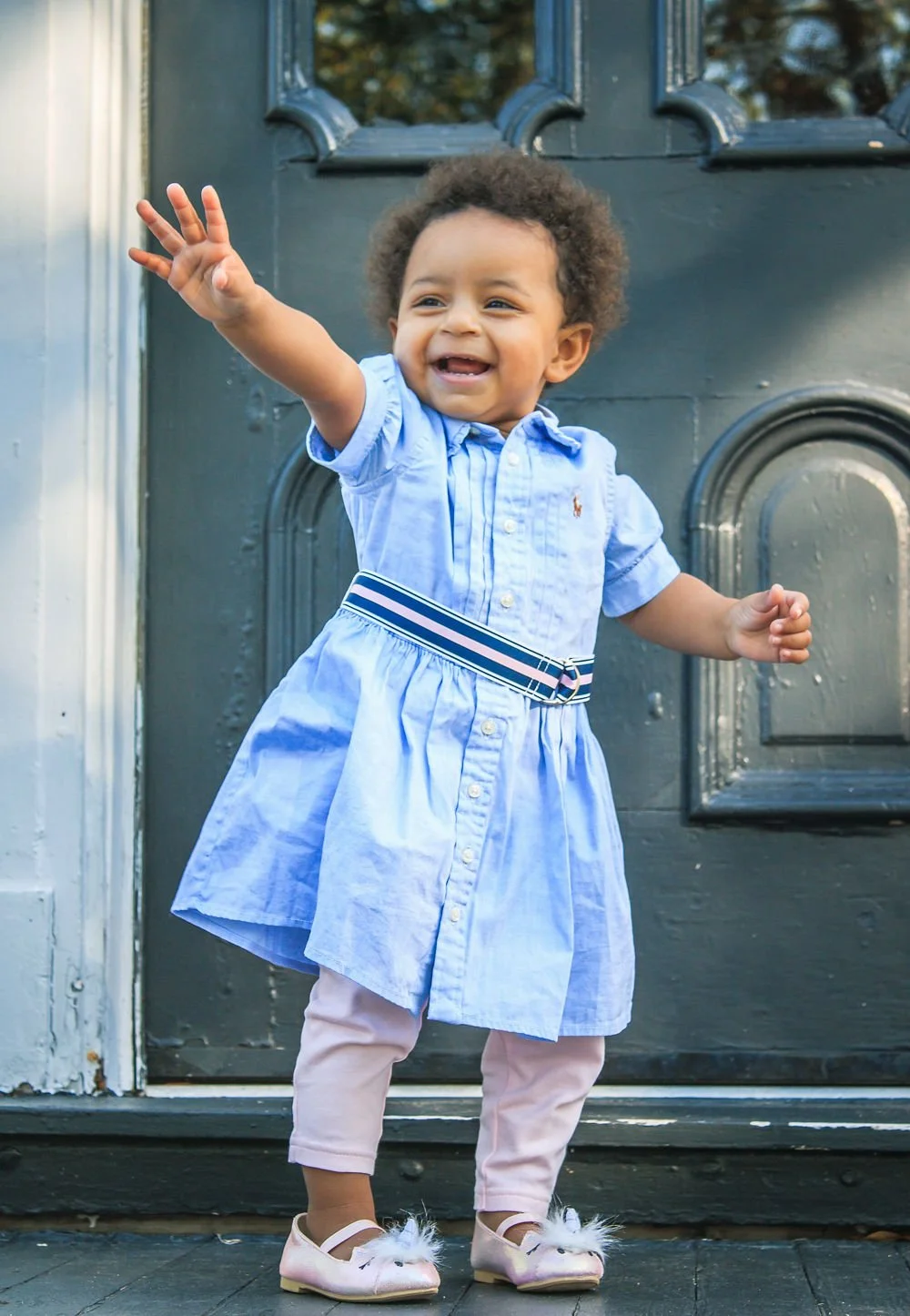 A young girl in a light blue dress, pink leggings, and pink shoes with white fur, smiling and waving in front of a black door.