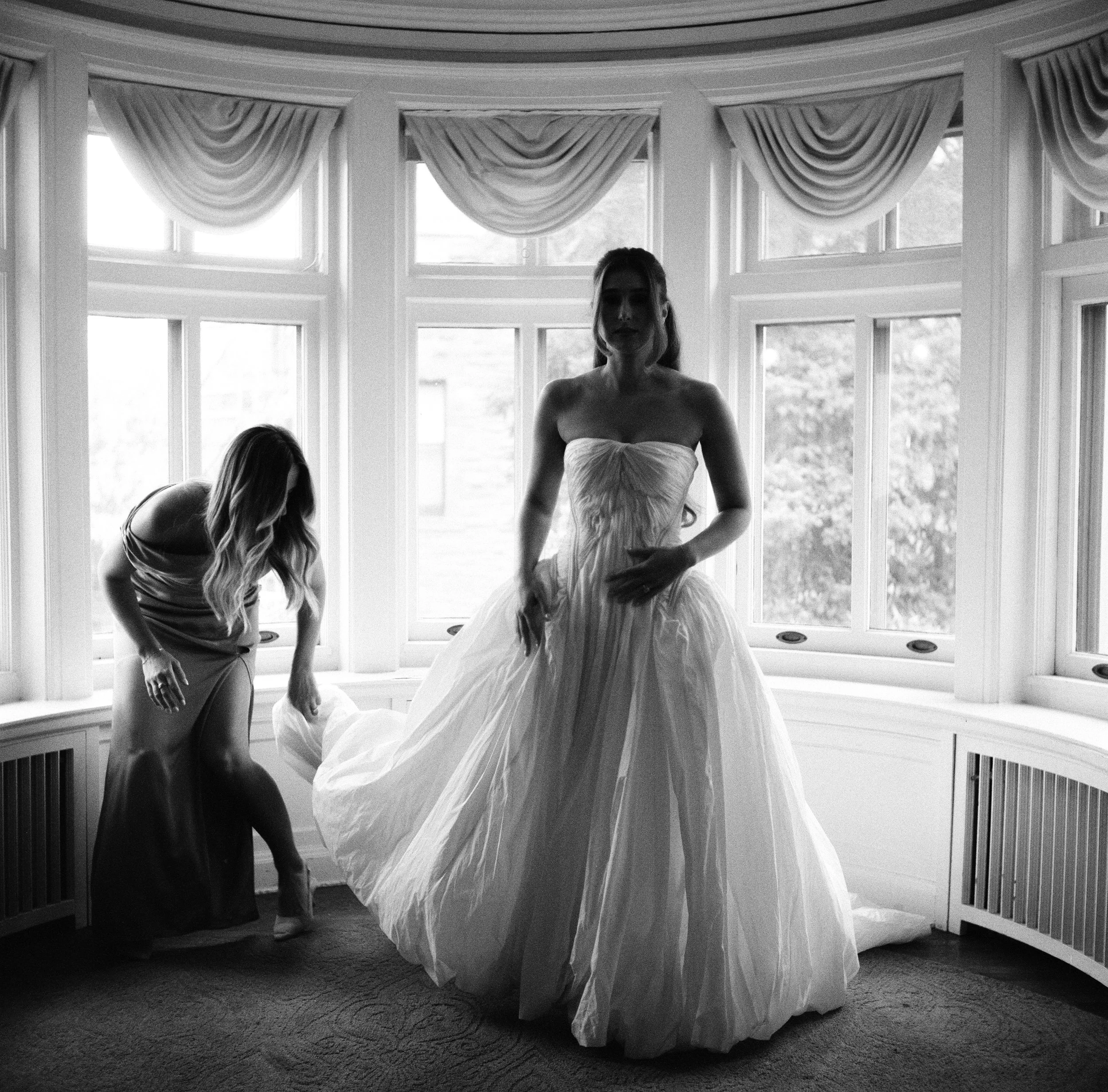 A woman in a wedding dress standing in front of large windows, with another woman adjusting her gown. The scene is in black and white.