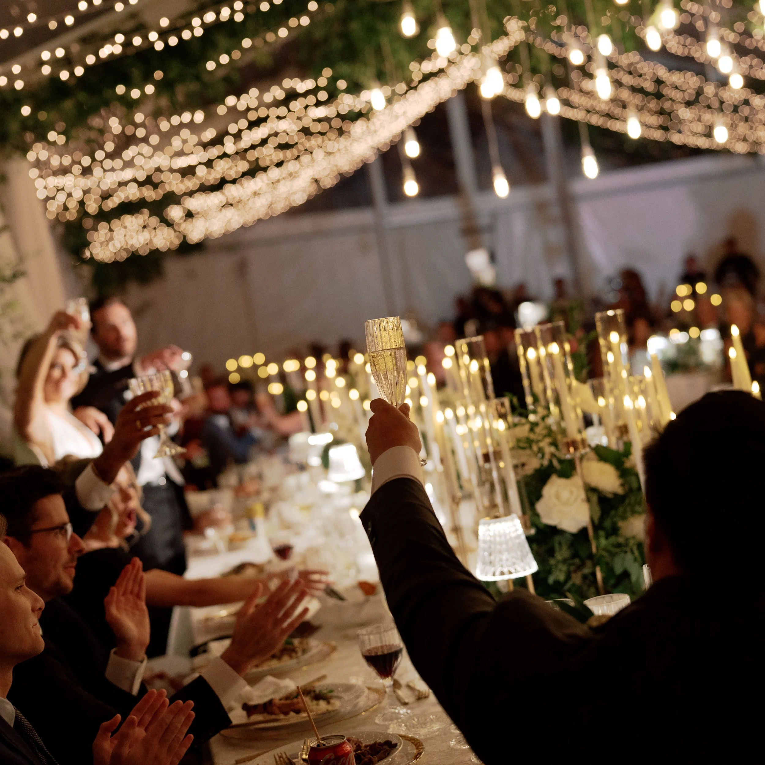 People raising glasses at a formal dinner table decorated with candles and flowers under string lights.