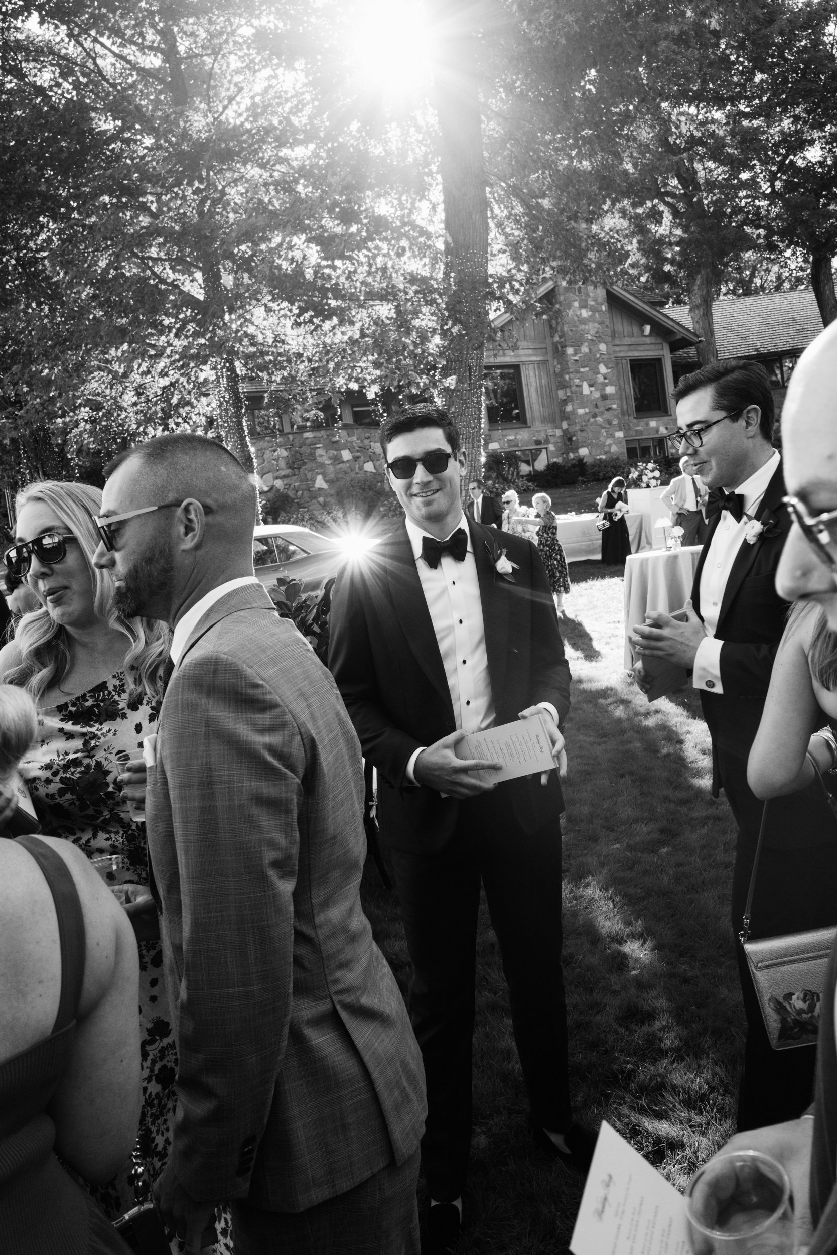 A black and white photo of a wedding reception outdoors with guests socializing. The central figure is a man in a tuxedo with sunglasses holding a program or menu, smiling. Other guests are dressed in formal attire, some wearing sunglasses, with a ba