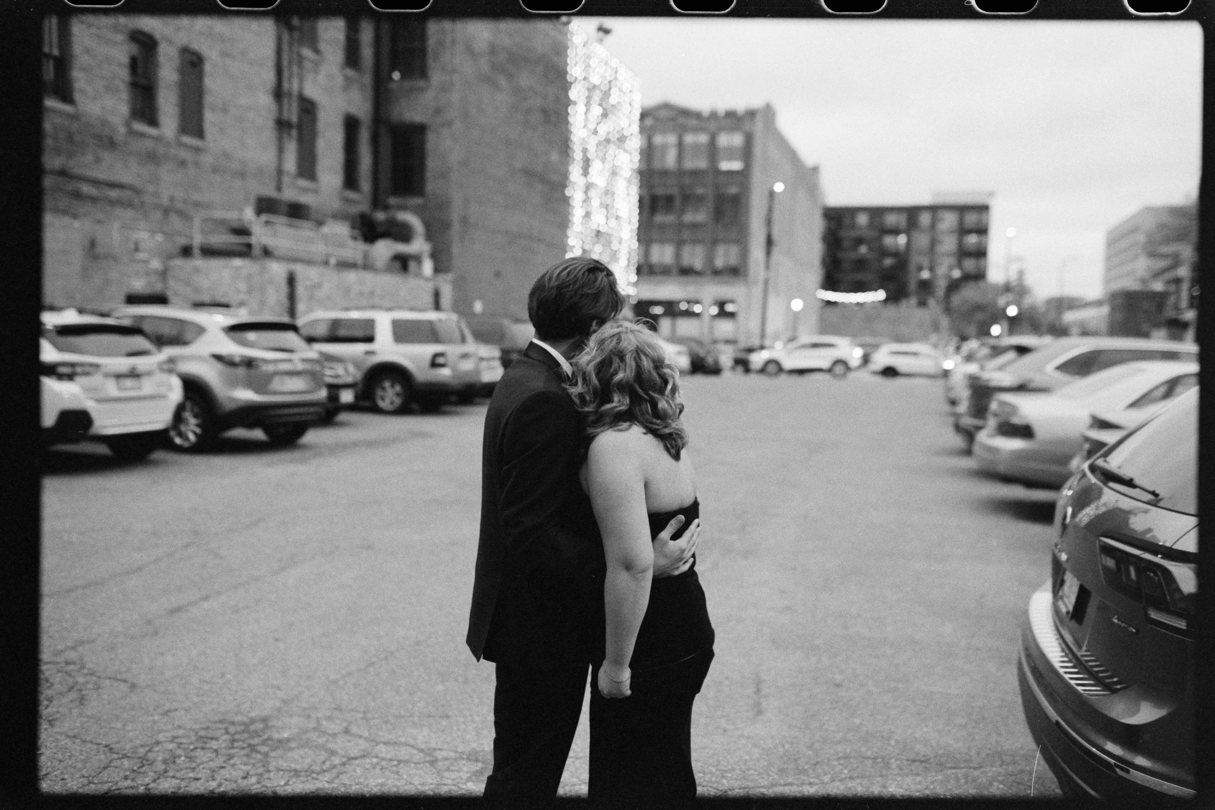 A black and white photo of a man and woman standing close together in a parking lot surrounded by cars, with city buildings in the background.