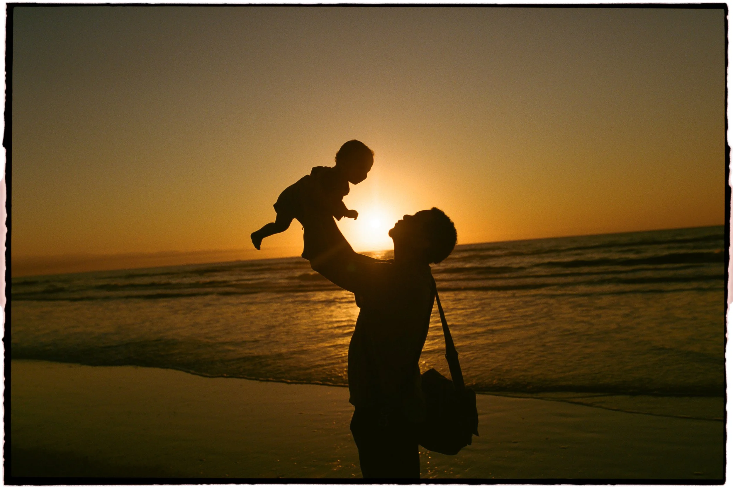 Silhouette of an adult and a child on the beach at sunset, with the child sitting on the adult's shoulders, against a backdrop of the ocean and a setting sun.