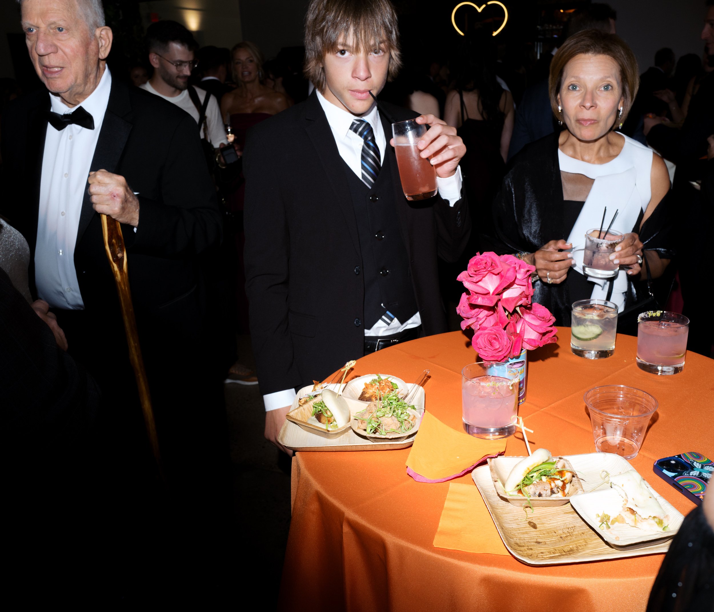 At a social gathering, a young man in a suit is drinking a pink beverage, standing near a table with pink flowers, tacos, and glasses of drinks, amidst other elegantly dressed guests.