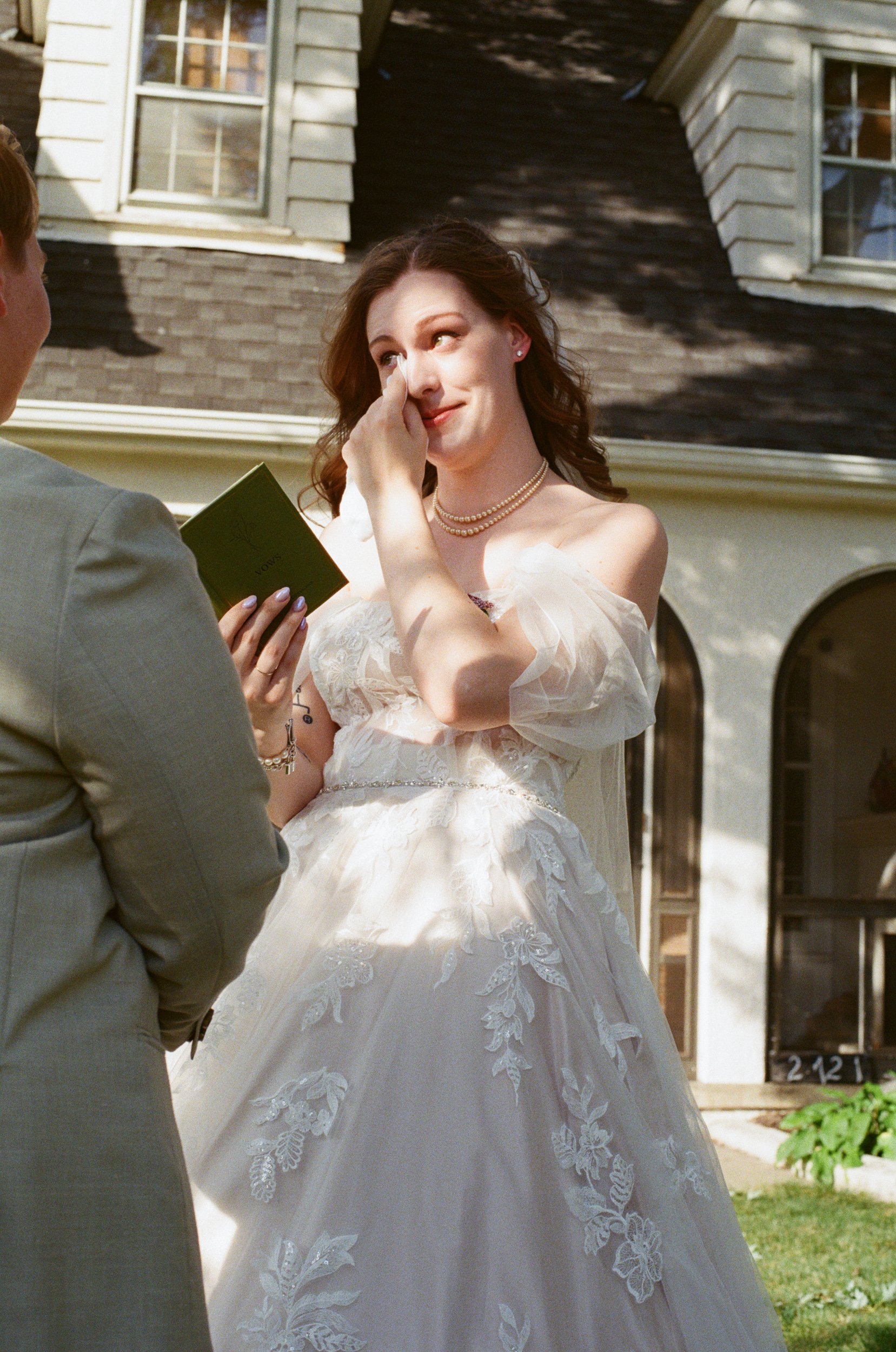 A bride in a white wedding dress wiping away tears during her wedding ceremony outside her house with a person holding a small green book.