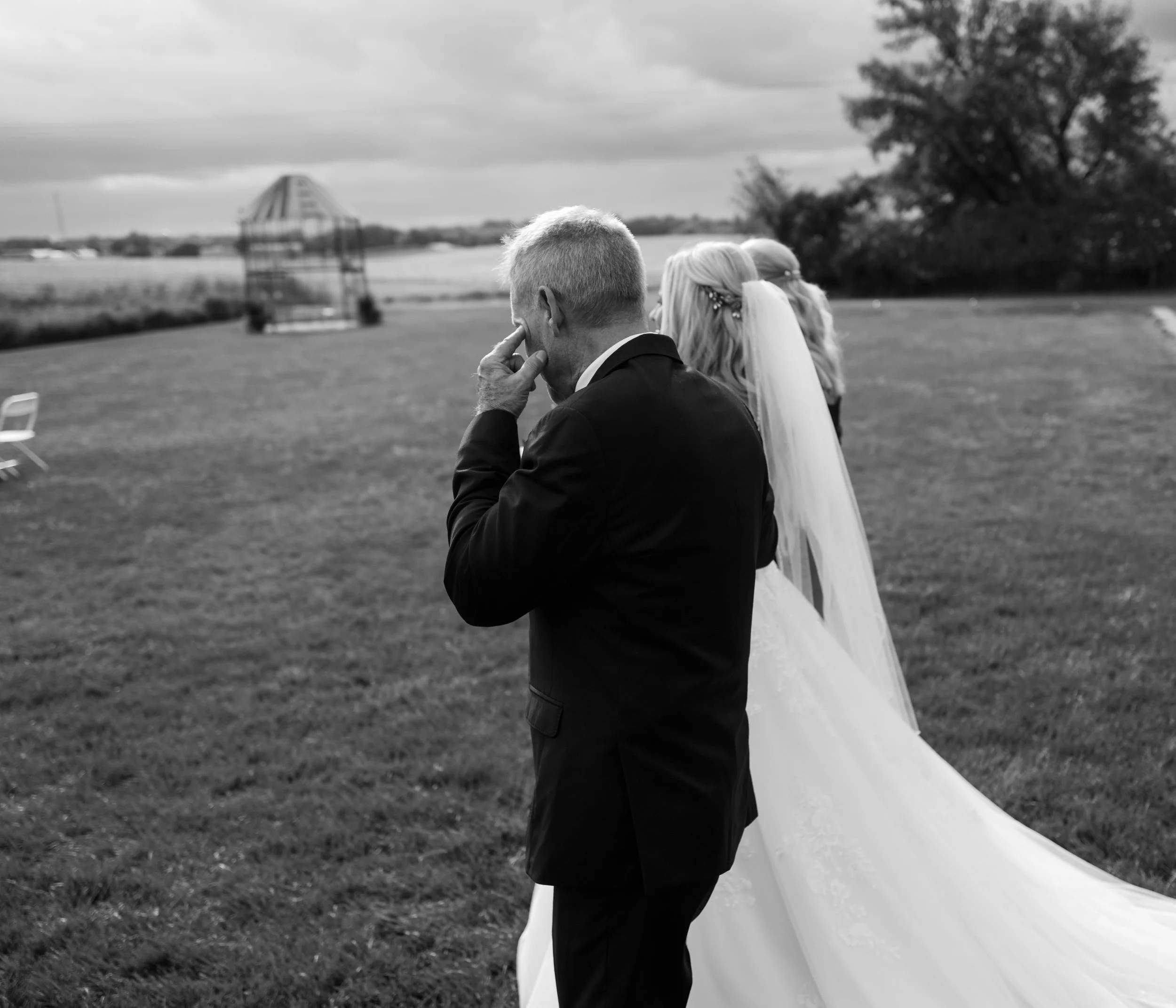 Black and white photo of a bride in a wedding dress and veil, standing beside a man in a suit, outdoors on grass with trees and a small structure in the background.