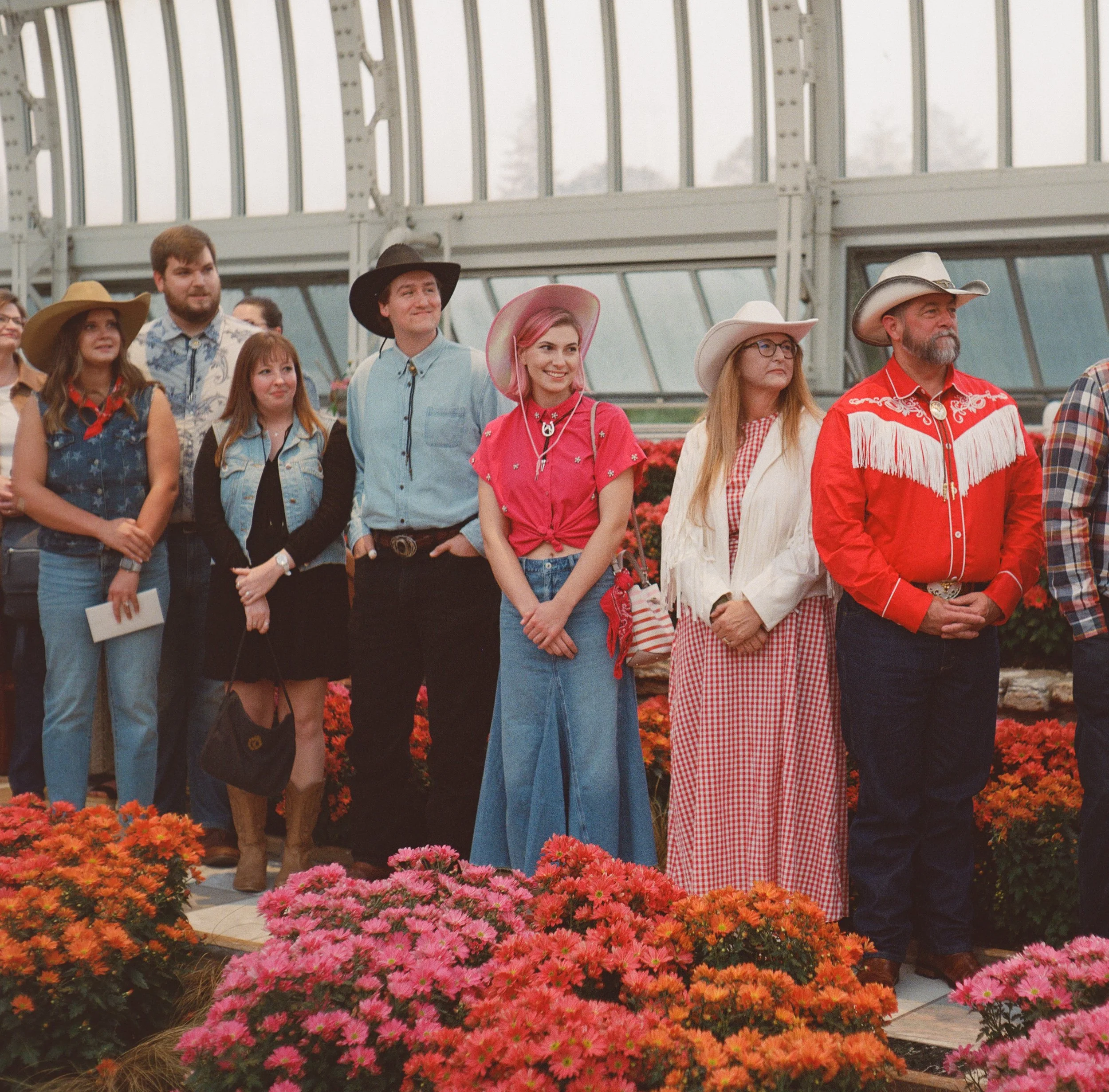 A group of people dressed in Western-style clothing, standing in a greenhouse surrounded by colorful flowers.