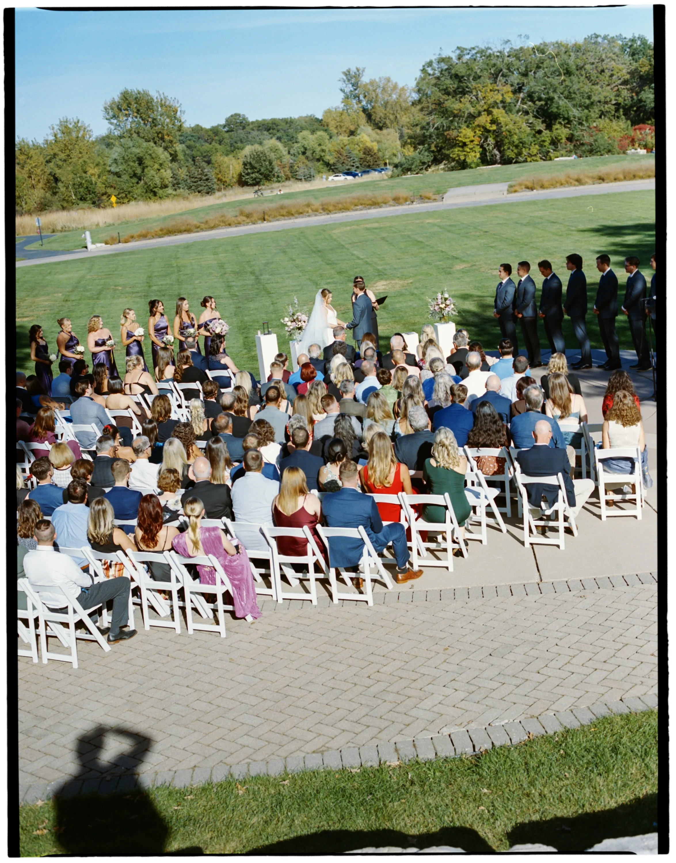 An outdoor wedding ceremony with a bride and groom standing in front of a officiant, surrounded by bridesmaids in purple dresses and groomsmen in suits. Guests are seated on white chairs. The setting is on a lush green lawn with trees and a parking lot in the background.