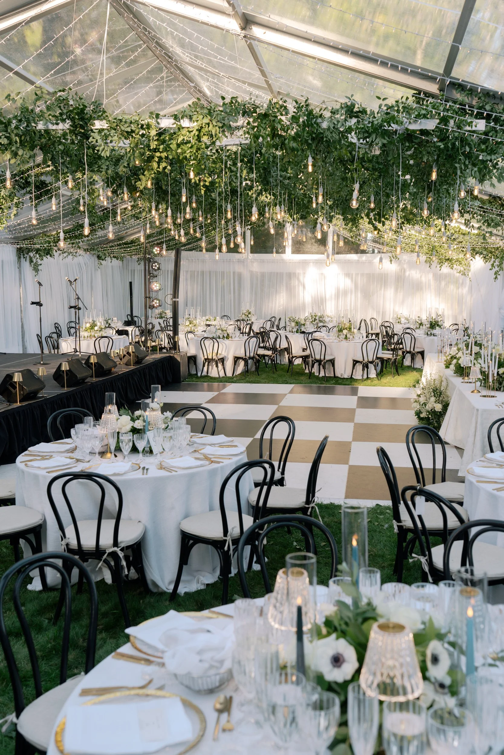 Elegant indoor wedding reception setup with round tables covered in white tablecloths, black chairs with white cushions, and floral centerpieces. Hanging string lights and greenery decorate the ceiling, with a live band stage at one end.