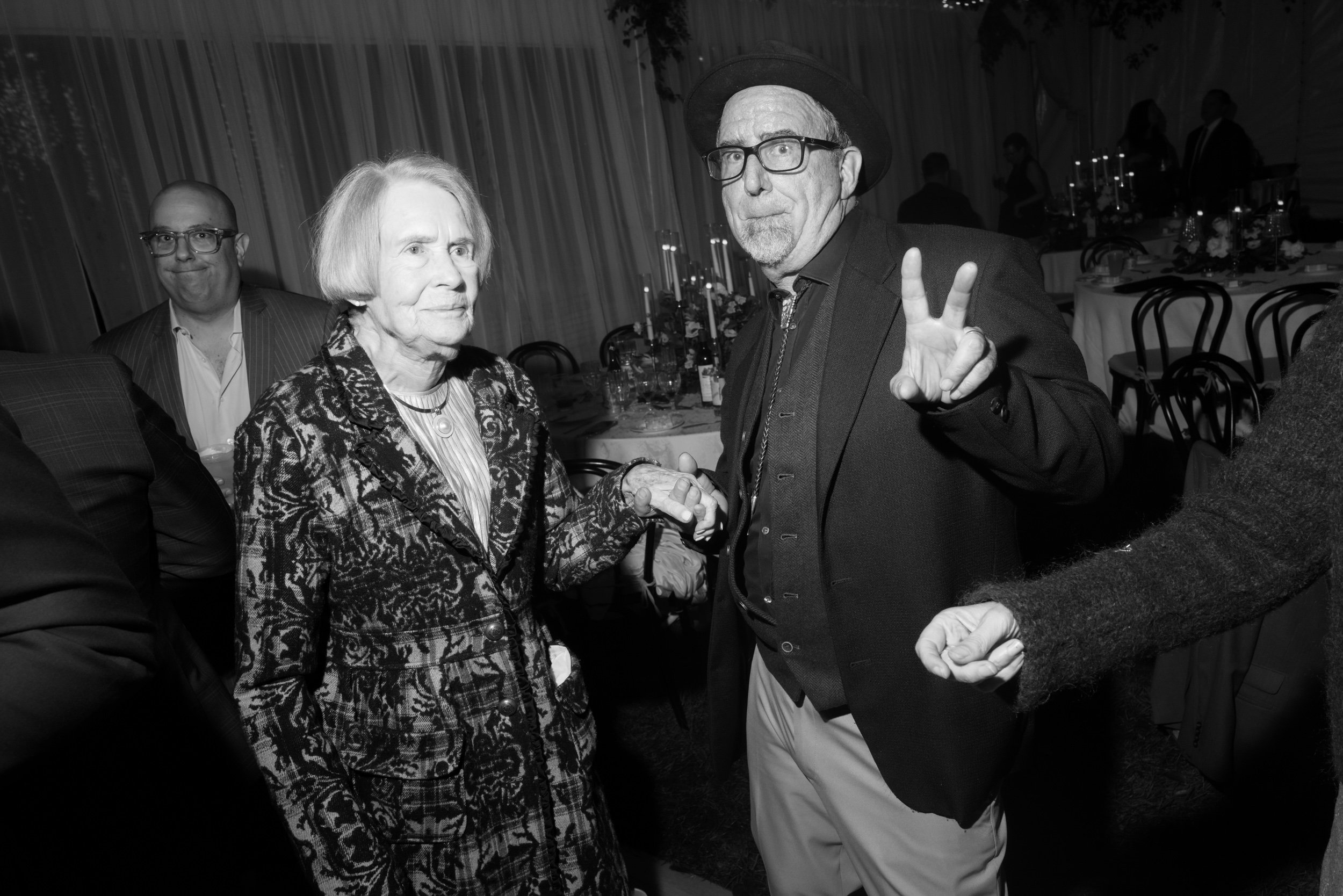 A black and white photo of several people at a social gathering or banquet. A man with glasses, a hat, and a dark blazer is making a peace sign gesture with his right hand and holding hands with an elderly woman. The elderly woman is wearing a patter