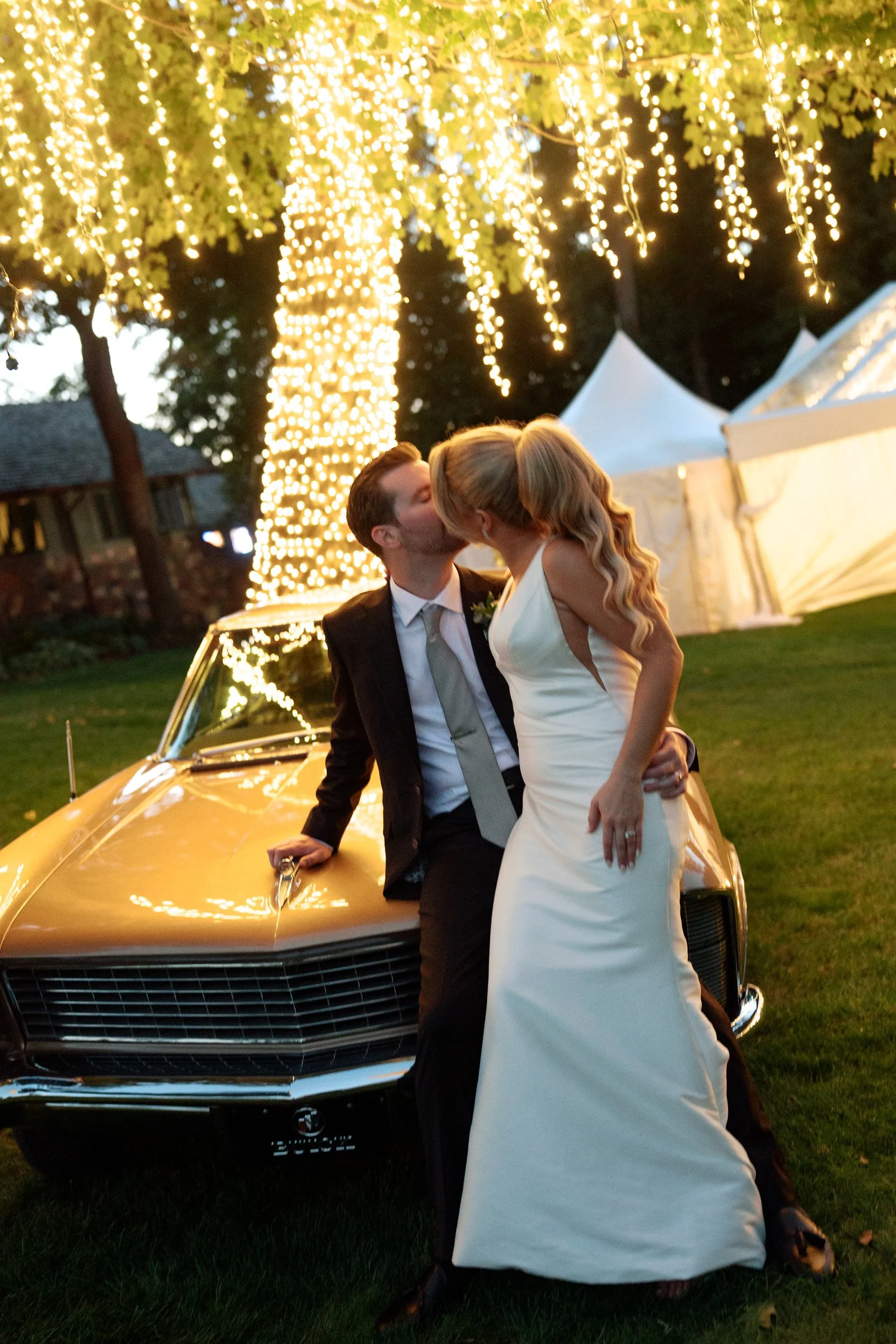 A newlywed couple sharing a kiss in front of a vintage car under a fairy light decorated tree at dusk.