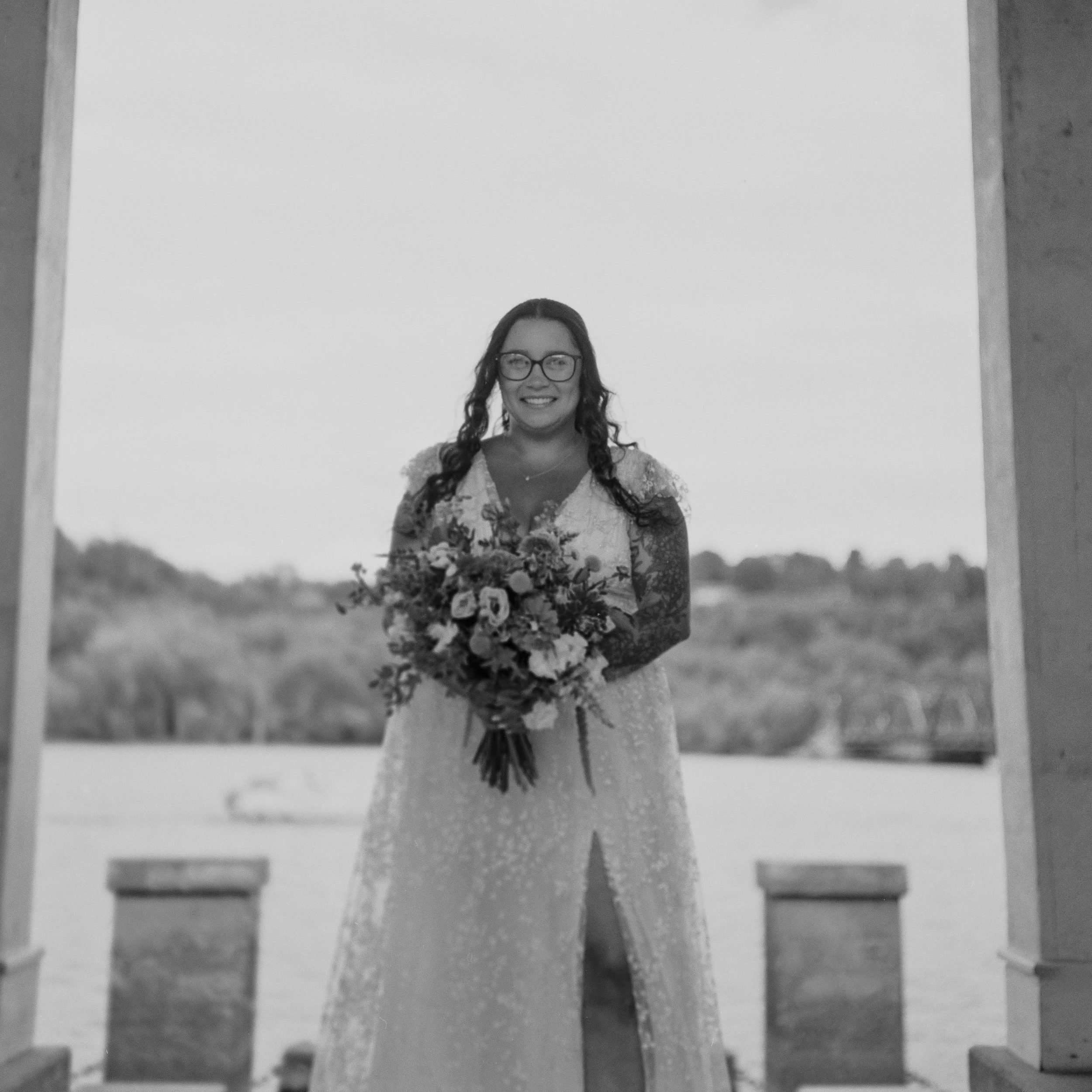A smiling woman in a wedding dress holding a bouquet, standing outdoors framed by a structure, with a scenic landscape in the background.