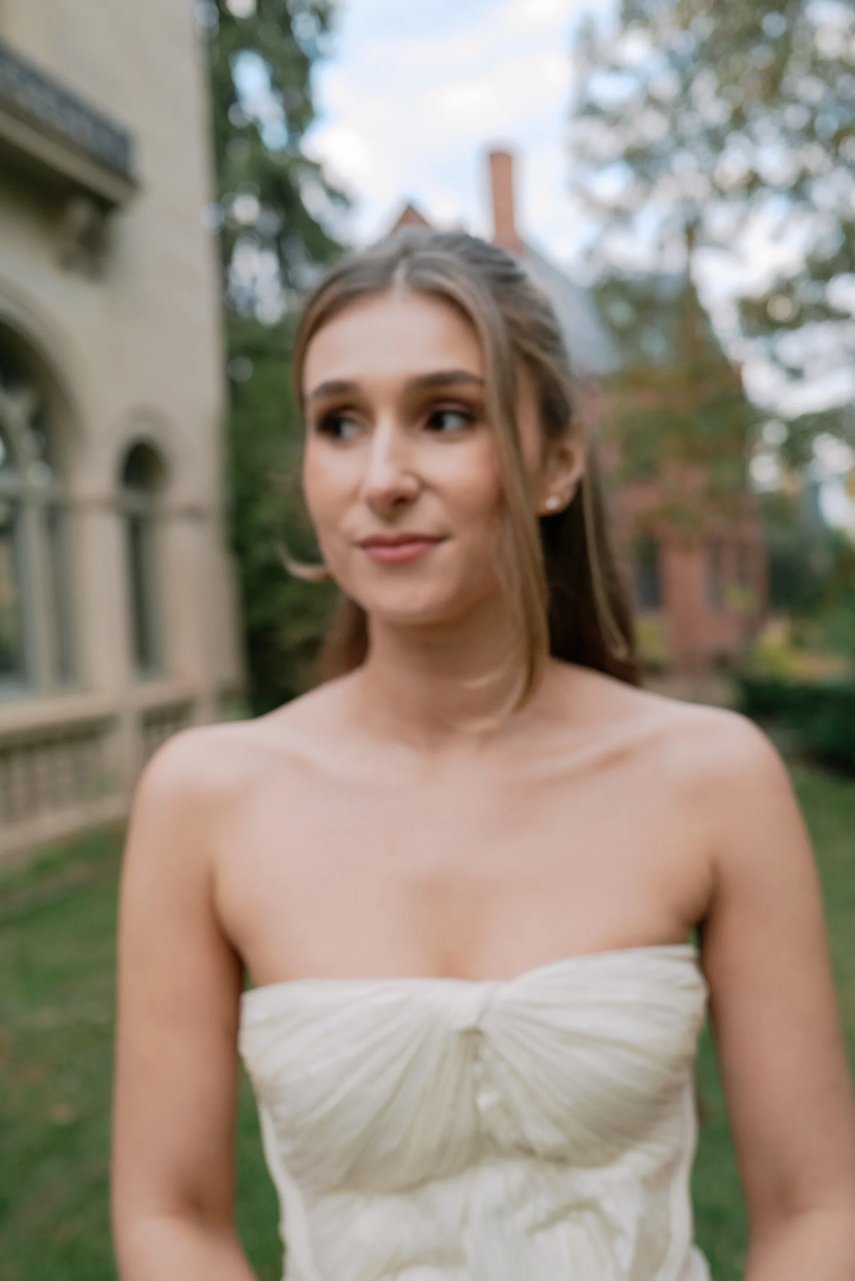 A young woman in a strapless cream-colored dress standing outdoors with a historic building and trees in the background.