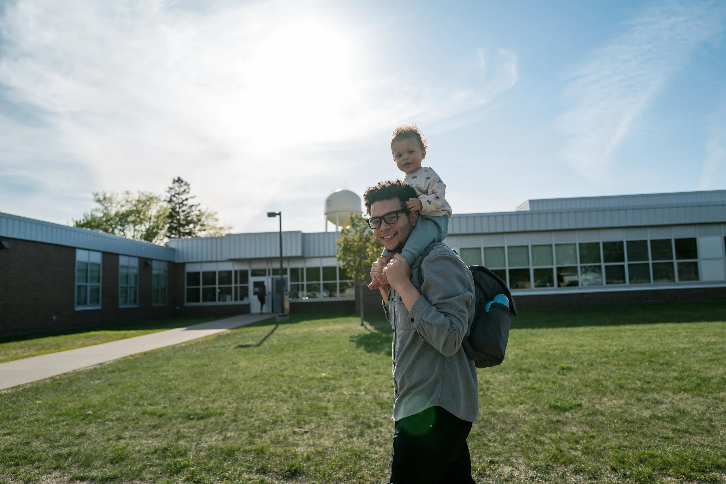A man with glasses carrying a young child on his shoulders outside a school building during daytime.