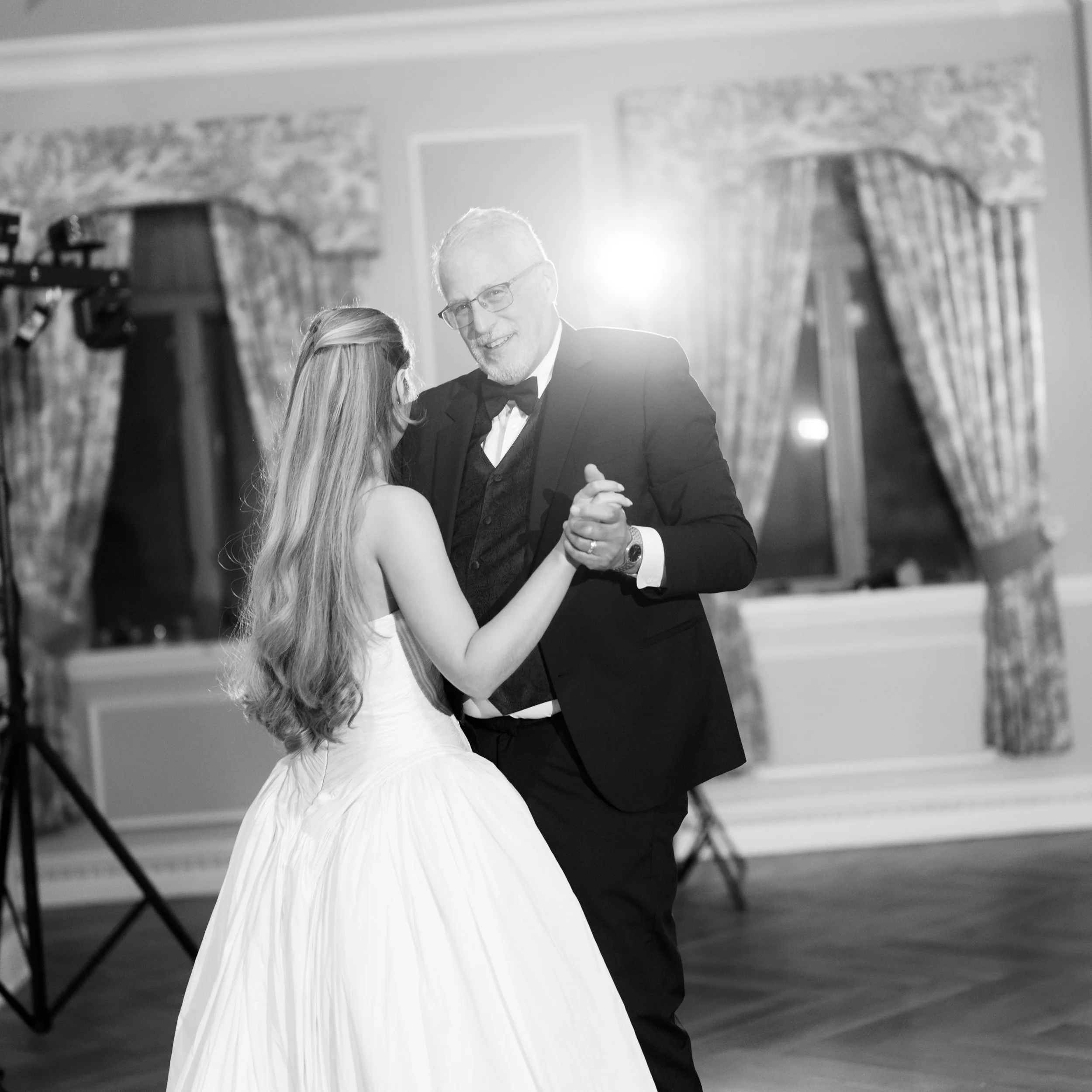 A man and young girl dancing at a formal event, with the girl wearing a white dress and the man in a tuxedo, in a decorated ballroom setting.
