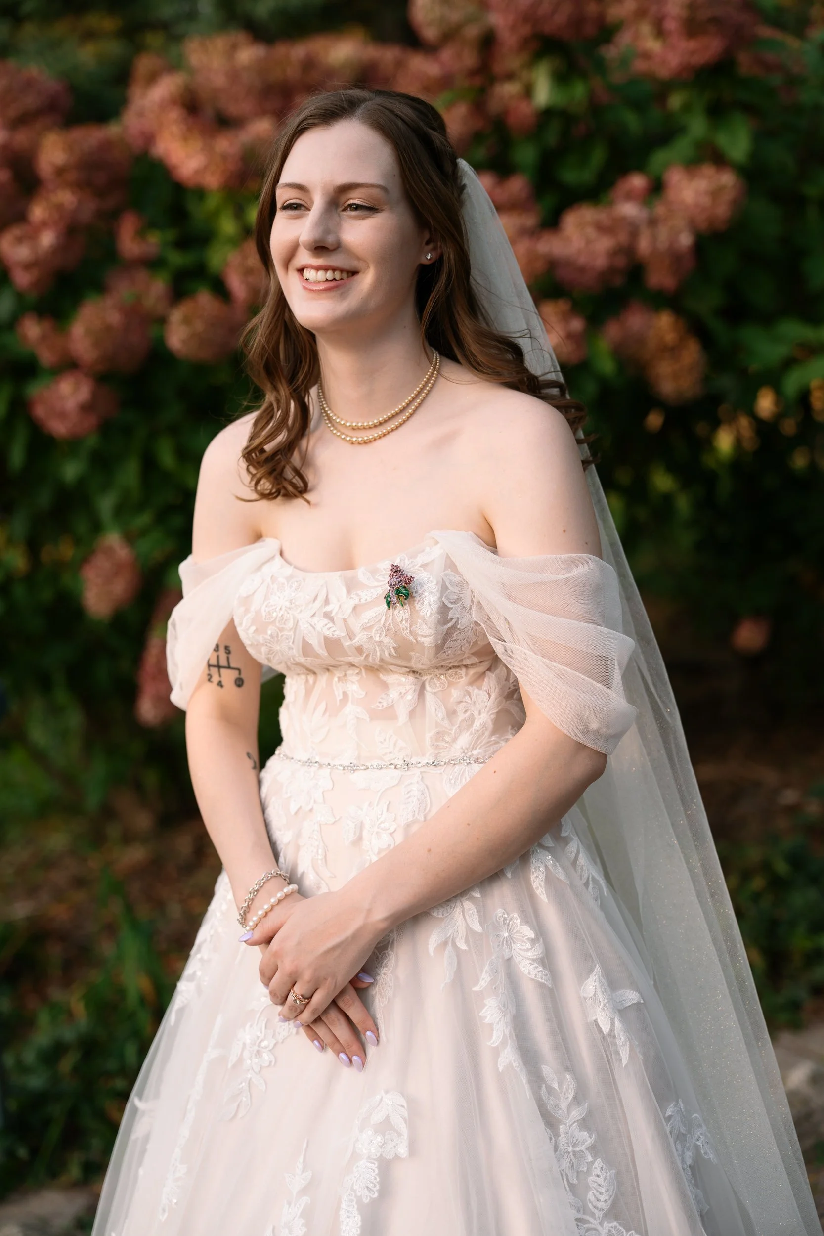 A smiling bride in a lace wedding dress with puffy sleeves, wearing pearl necklaces and a veil, standing outdoors in front of pink hydrangea flowers.