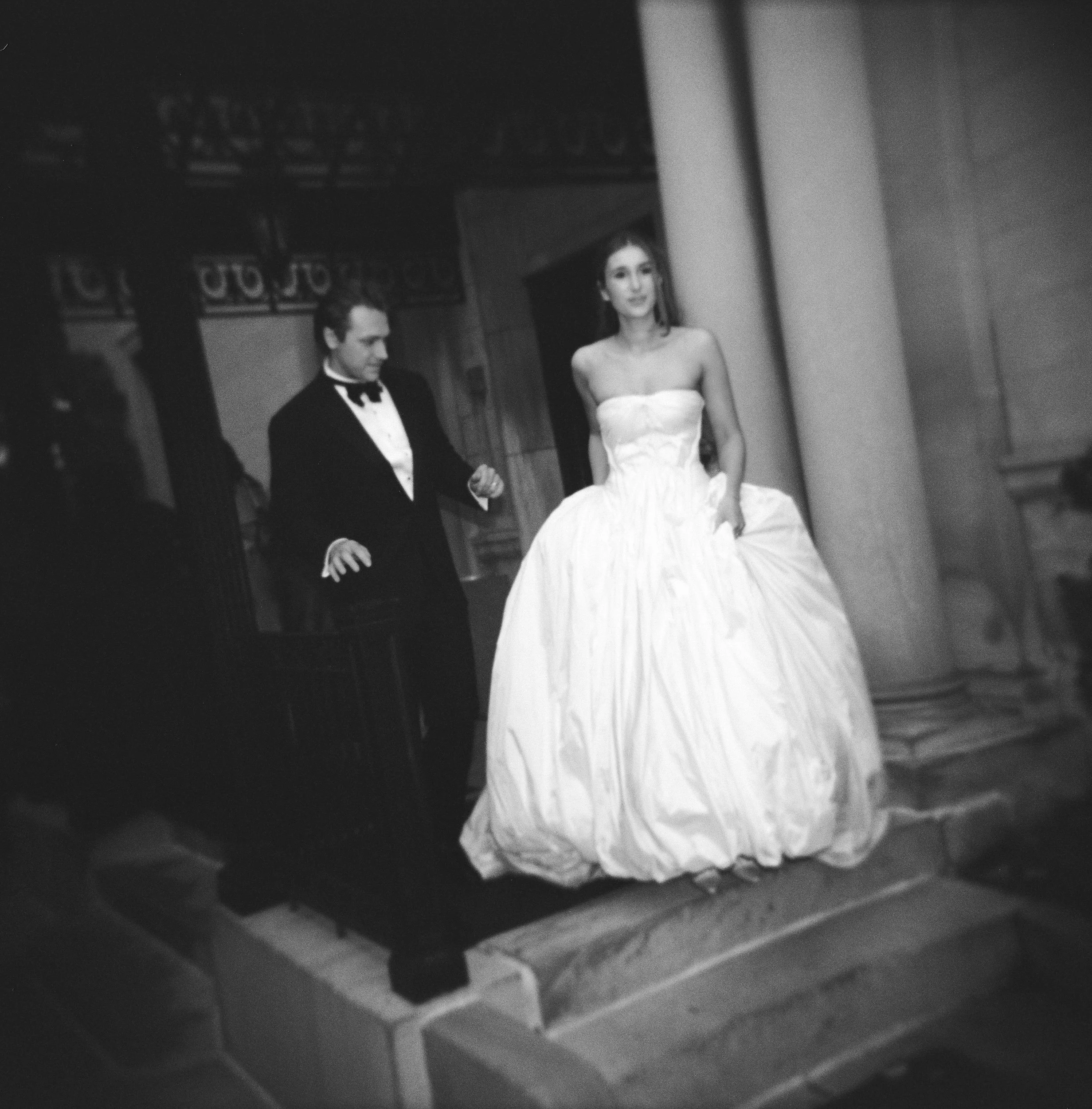 A black and white photo of a bride and groom standing on a staircase inside a building or church. The bride is wearing a strapless wedding gown, and the groom is dressed in a tuxedo. The bride is looking forward, while the groom is looking down.