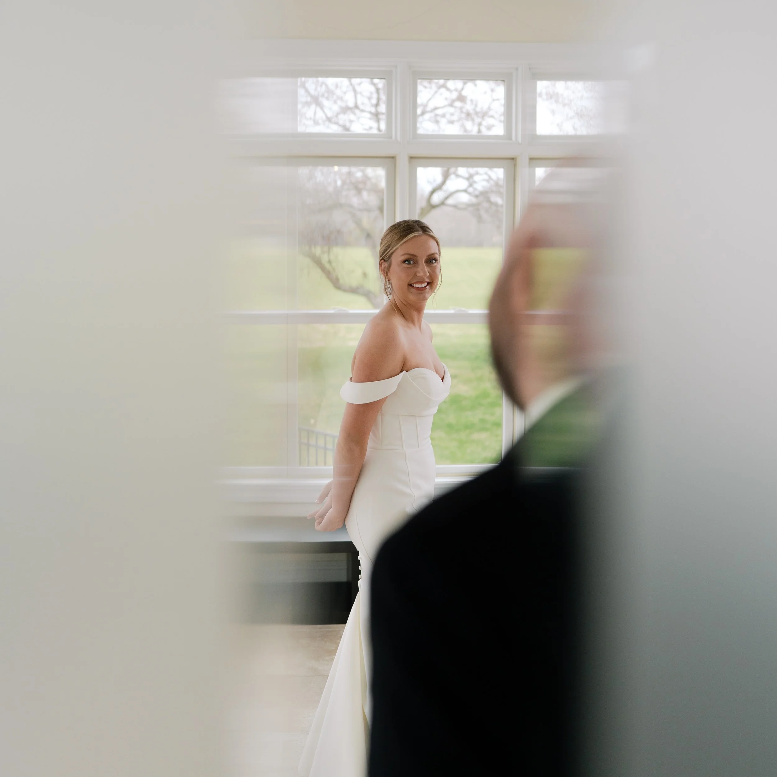 A bride in a white off-the-shoulder wedding gown smiling as she looks toward a man in a tuxedo, with a large window showing a green outdoor landscape in the background.