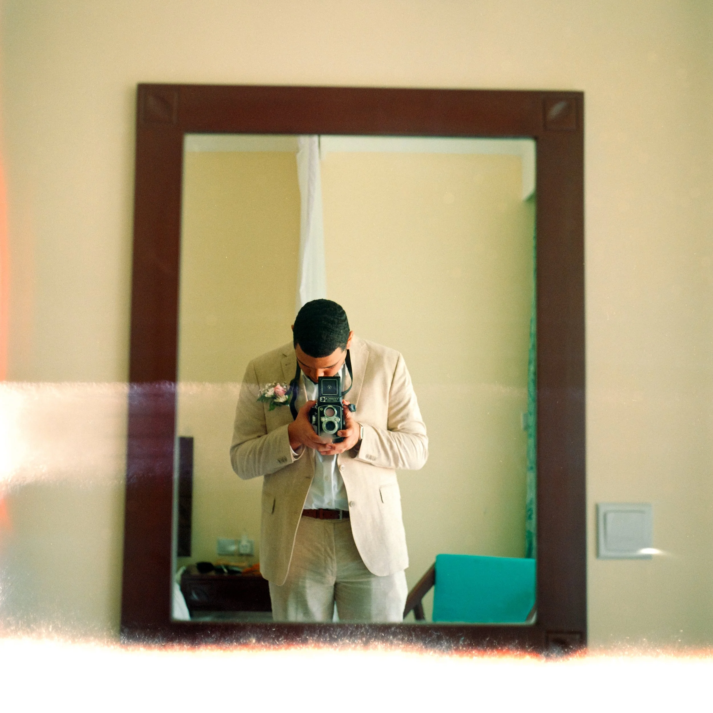 A person dressed in a light-colored suit taking a selfie in a rectangular mirror with a dark wooden frame, in a room with green walls and various furniture and objects.