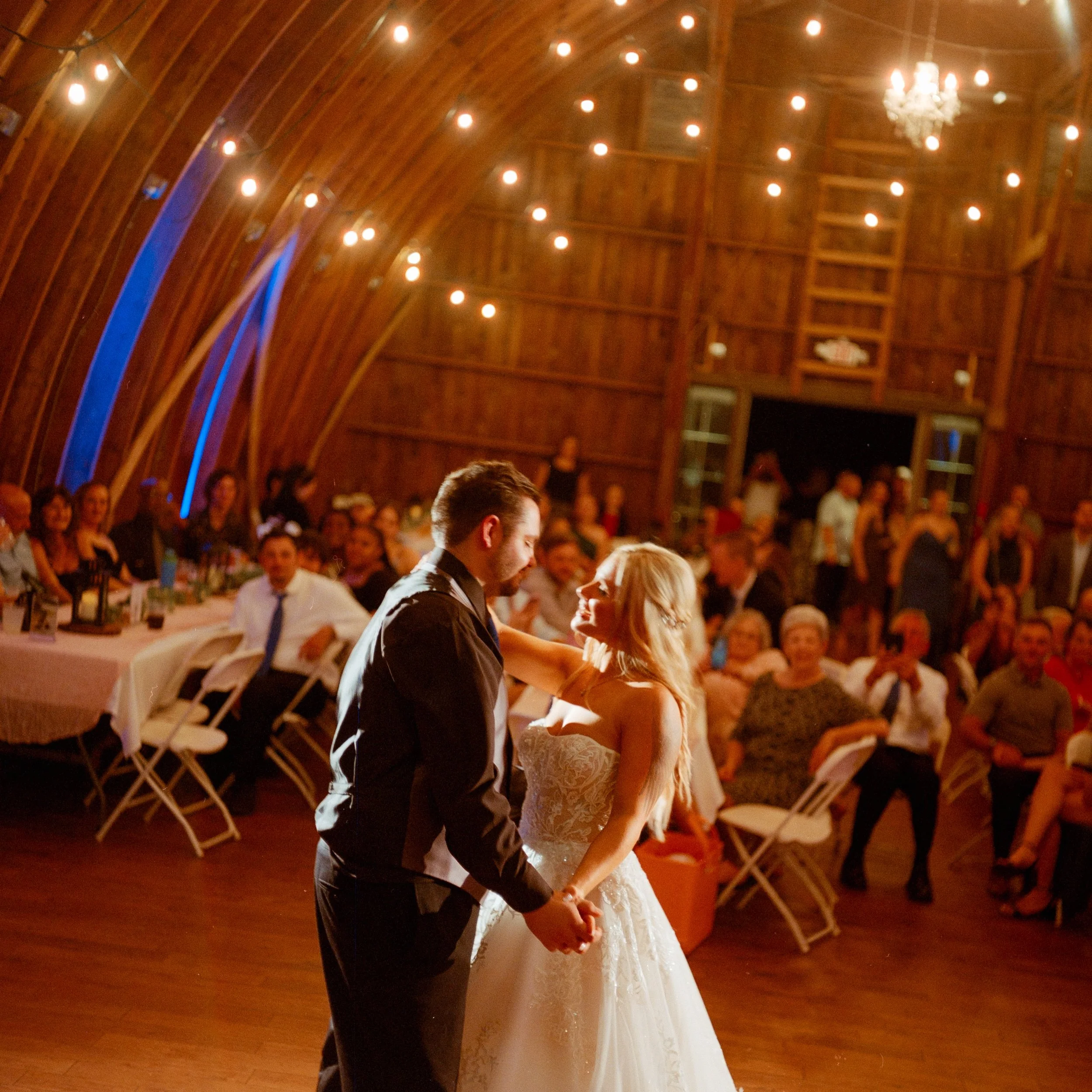 A bride and groom dance together at their wedding reception inside a wooden barn with string lights and a chandelier, surrounded by seated guests.