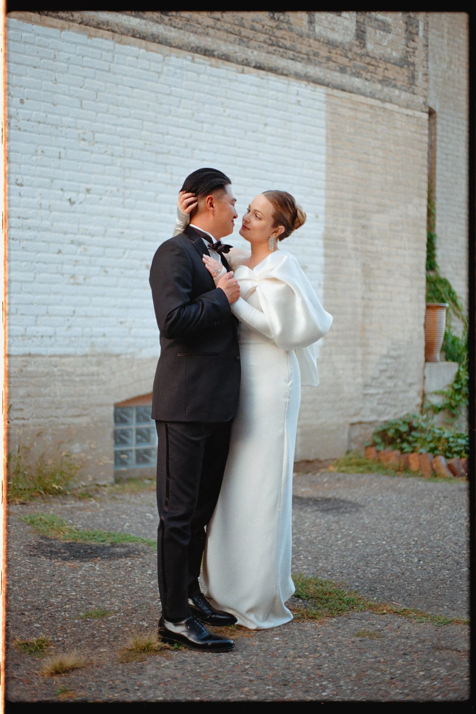 A bride and groom in wedding attire sharing a romantic moment outdoors against a brick wall.