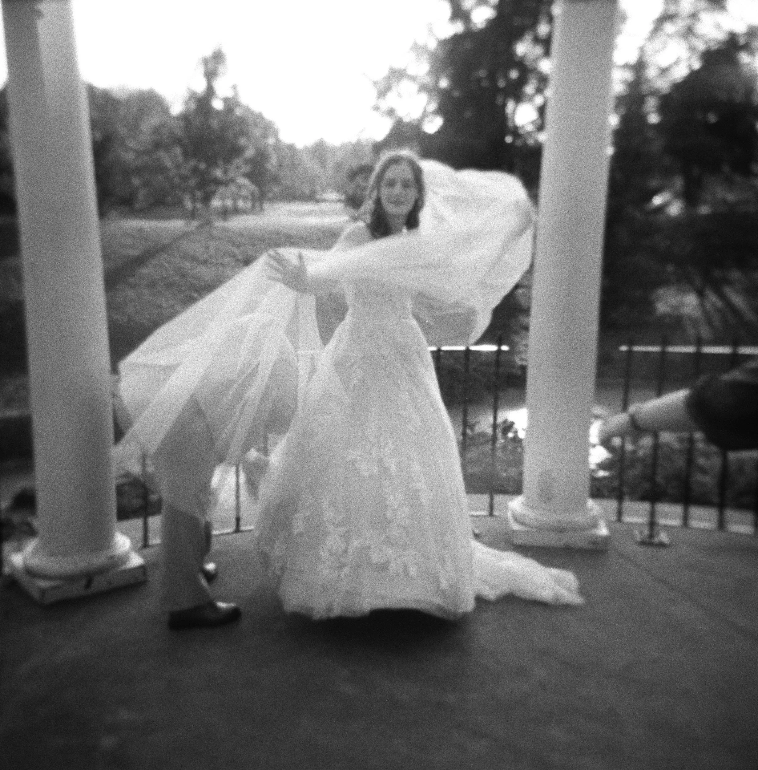 A bride in a wedding gown and a young boy in formal clothing on a porch with columns, with trees, grass, and a fence in the background.
