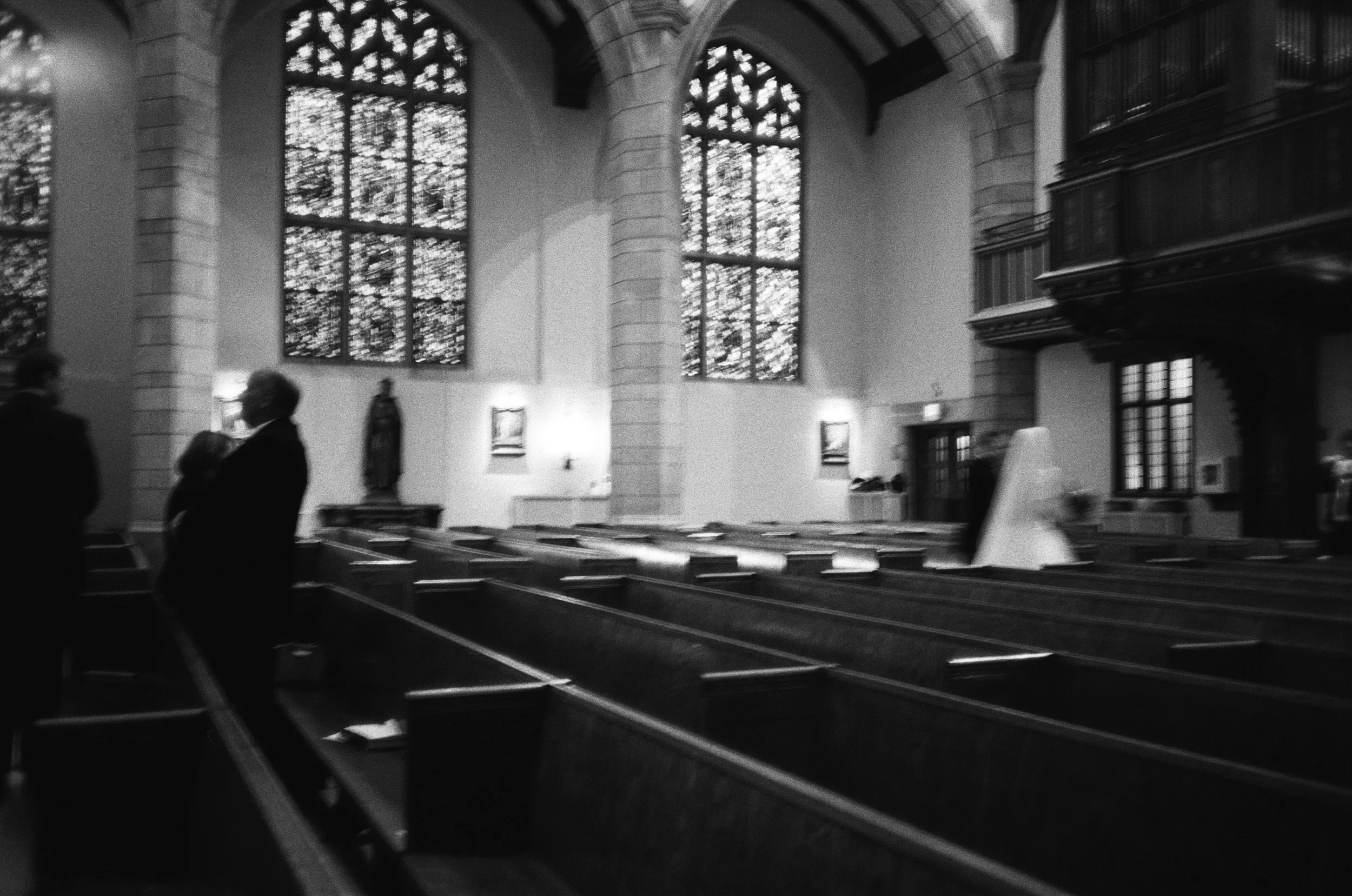 Inside a church with large stained glass windows, wooden pews, and a few people standing and talking.