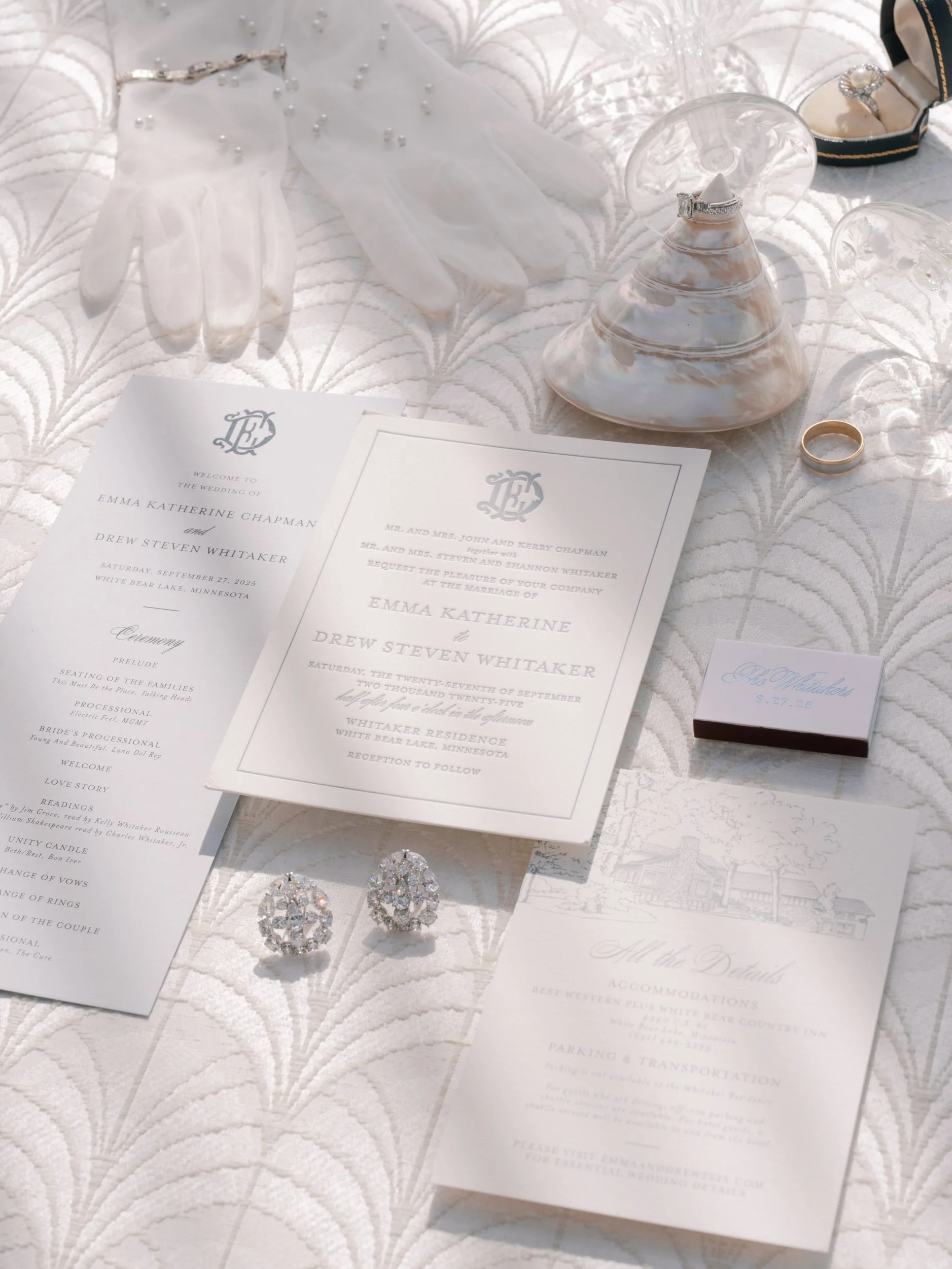 Wedding invitation suite with rings, jewelry, seashells, gloves, and wedding details on a white textured fabric.