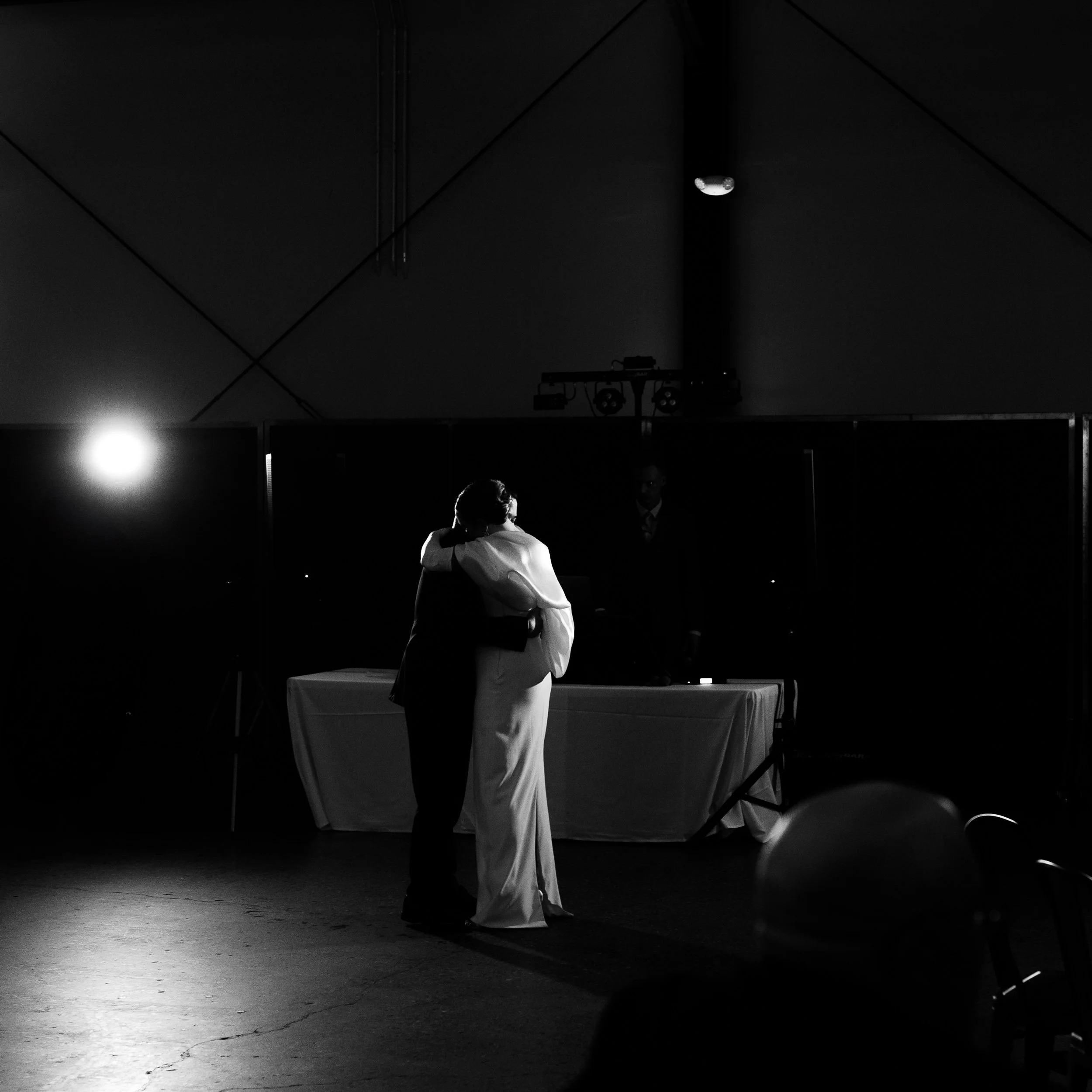 A couple in wedding attire embraces on a dance floor during a wedding reception with a DJ in the background.