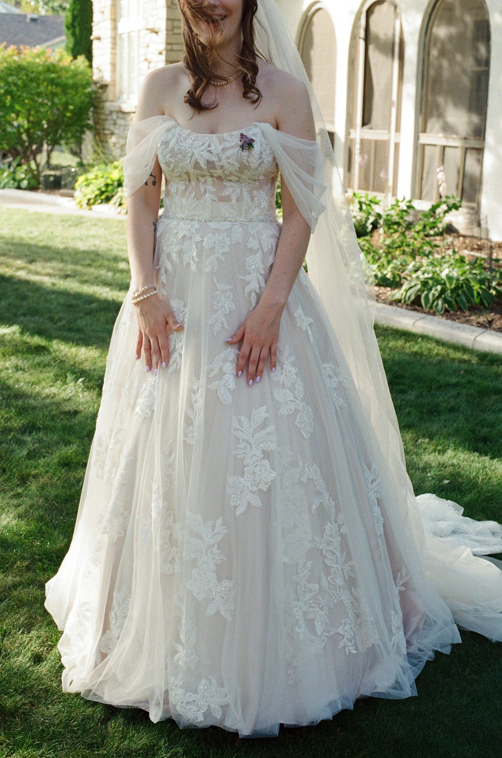 A bride wearing a white wedding dress with floral embroidery, standing outdoors on a grassy lawn. She is smiling, with her hands resting on her dress, and is wearing jewelry including a pearl bracelet and a necklace. She has loose hair with a veil at