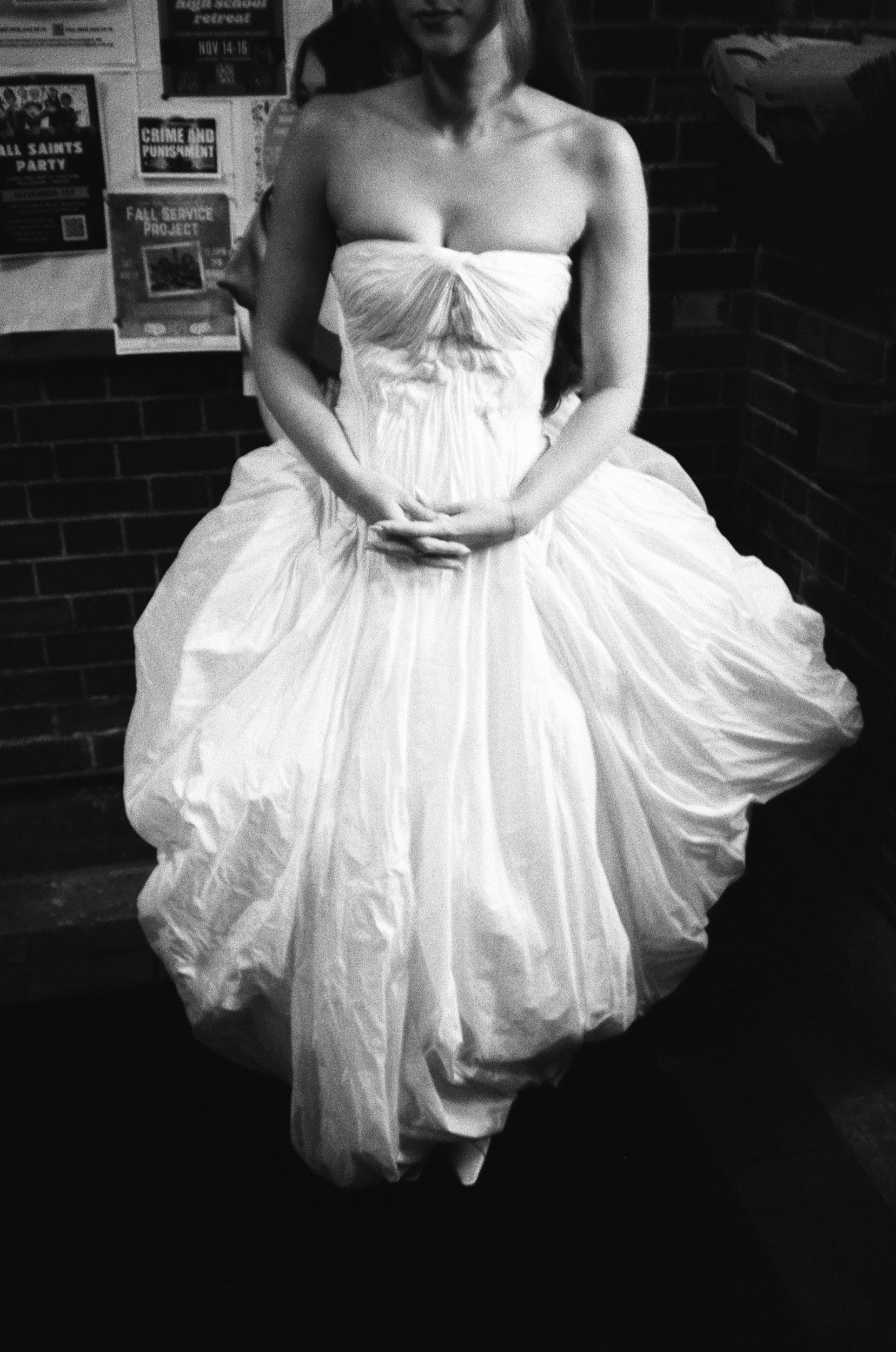 A woman in a strapless white wedding dress sitting on a bench in a brick-walled room, with posters on the wall behind her.