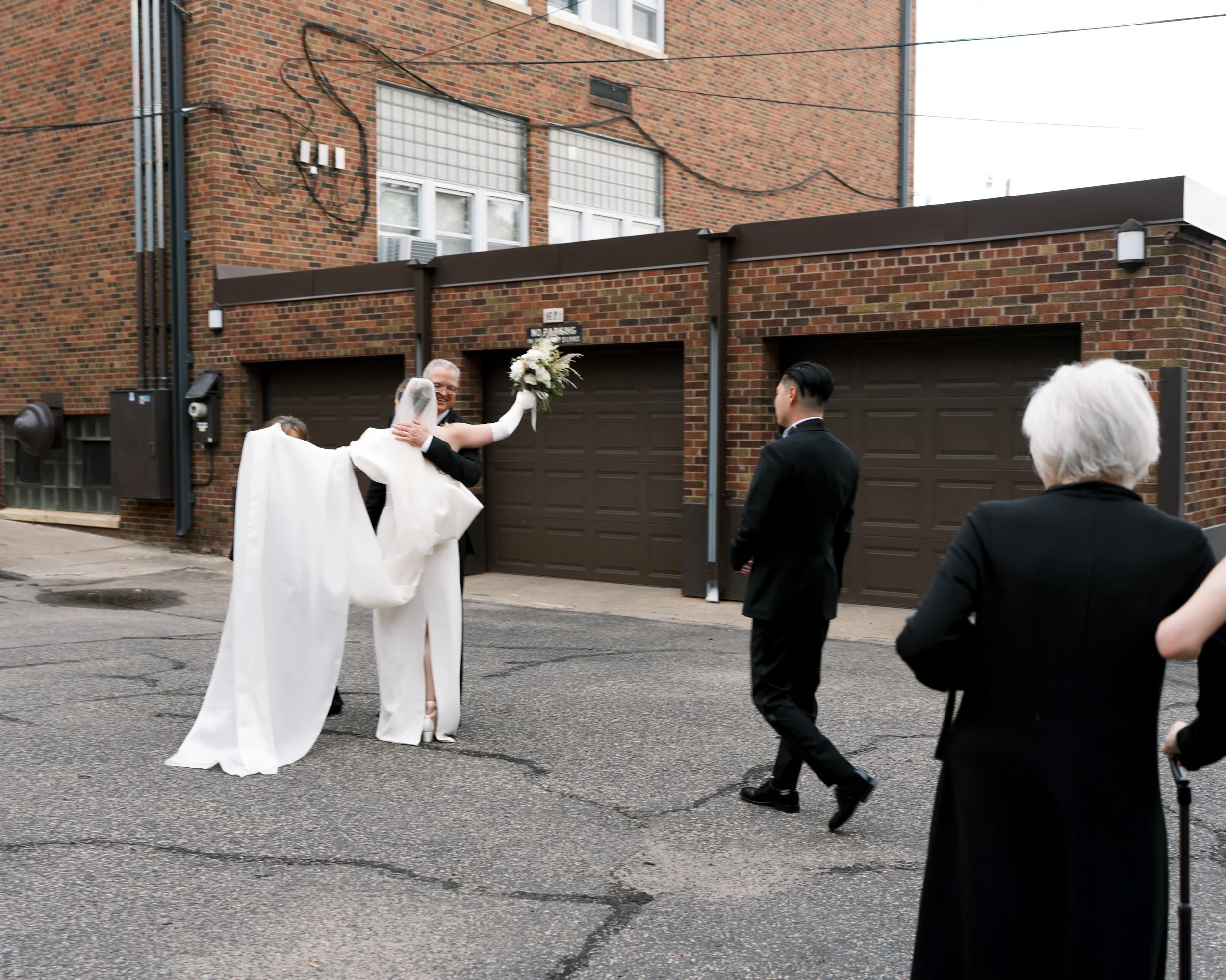 A bride in a white wedding dress and veil is being lifted by her groom in a black suit, smiling and holding a bouquet, on a city street with three other people in black formal attire watching, against a background of brick buildings and garage doors.