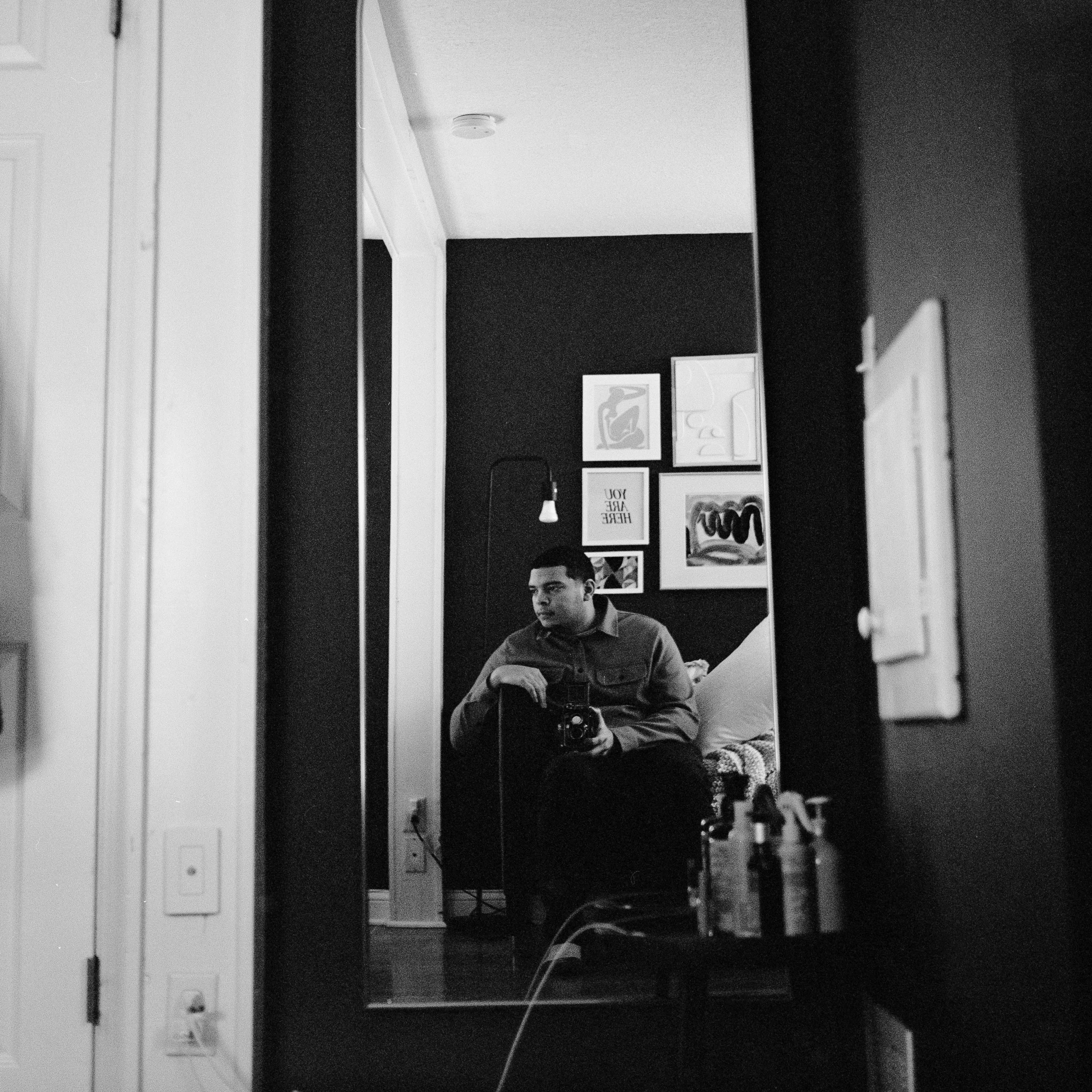 A person sitting in front of a mirror holding a camera, taking a selfie in a room with framed artwork on the wall.