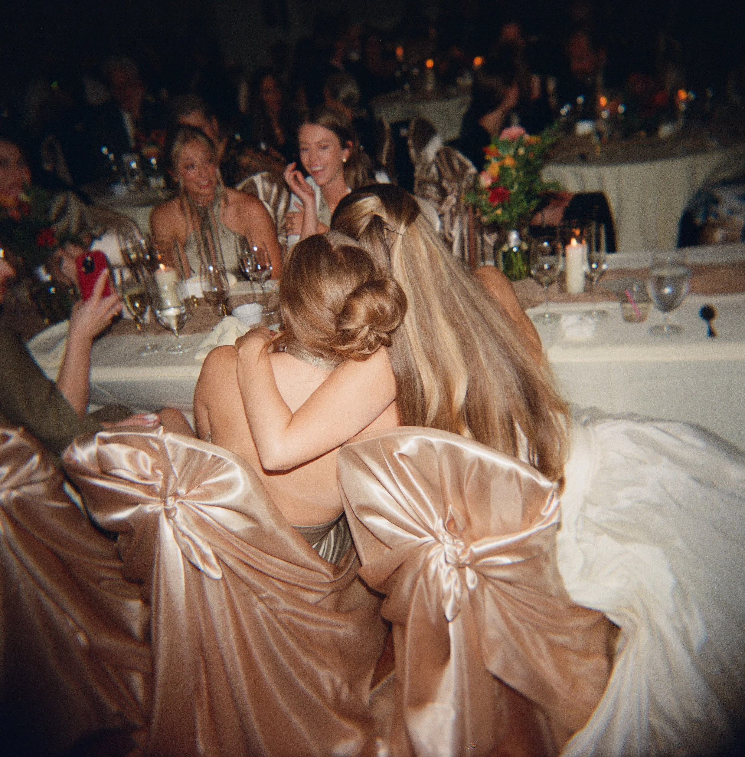 Two women hugging each other at a wedding reception with other guests seated at tables in the background.