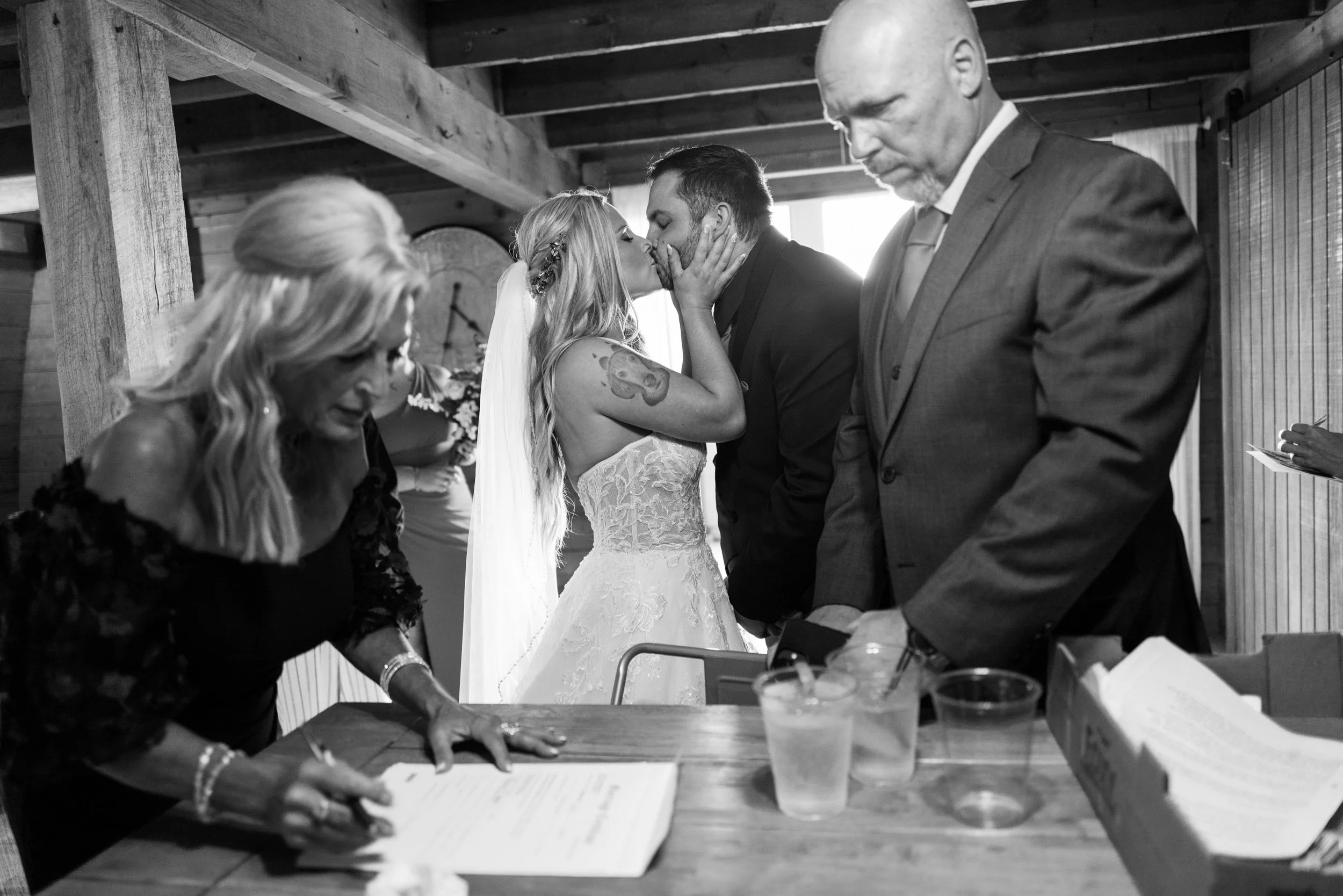 A bride and groom share a kiss during their wedding ceremony, with witnesses and an officiant present in a rustic indoor setting.
