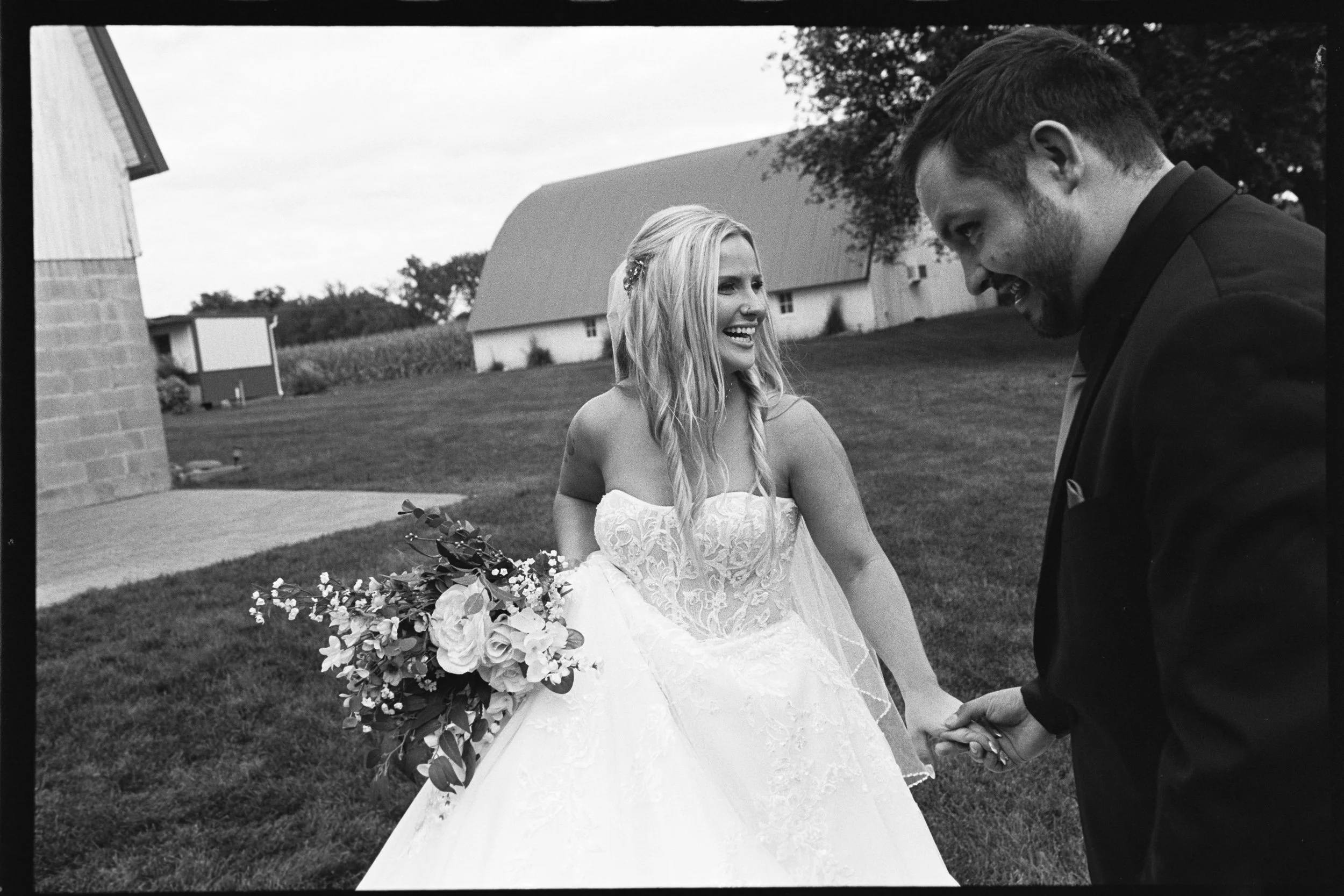 A bride and groom smiling and holding hands outdoors during a wedding celebration, with the bride wearing a strapless wedding dress and holding a bouquet, and the groom dressed in a suit.