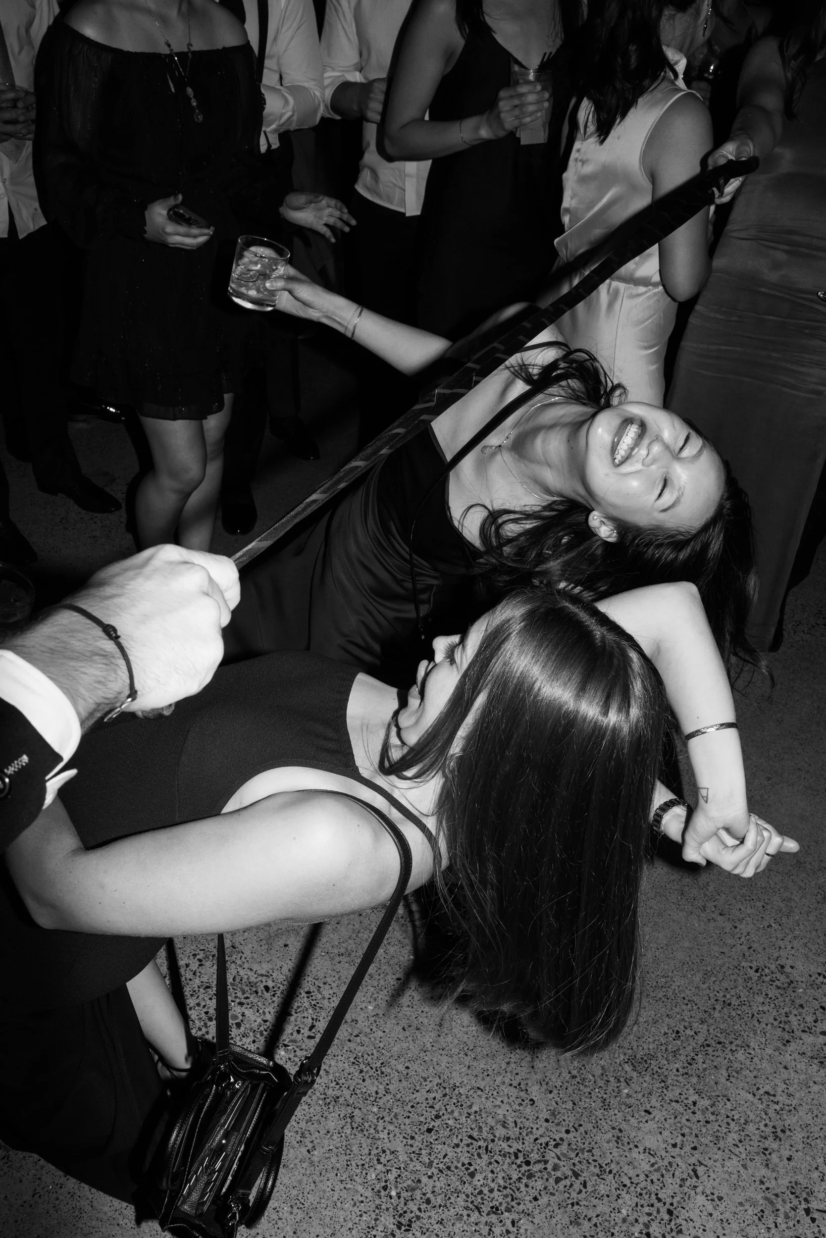 Two women are dancing together at a party, laughing and having fun, while a hand holds a drink in the foreground. Other people are seen in the background holding drinks and socializing.