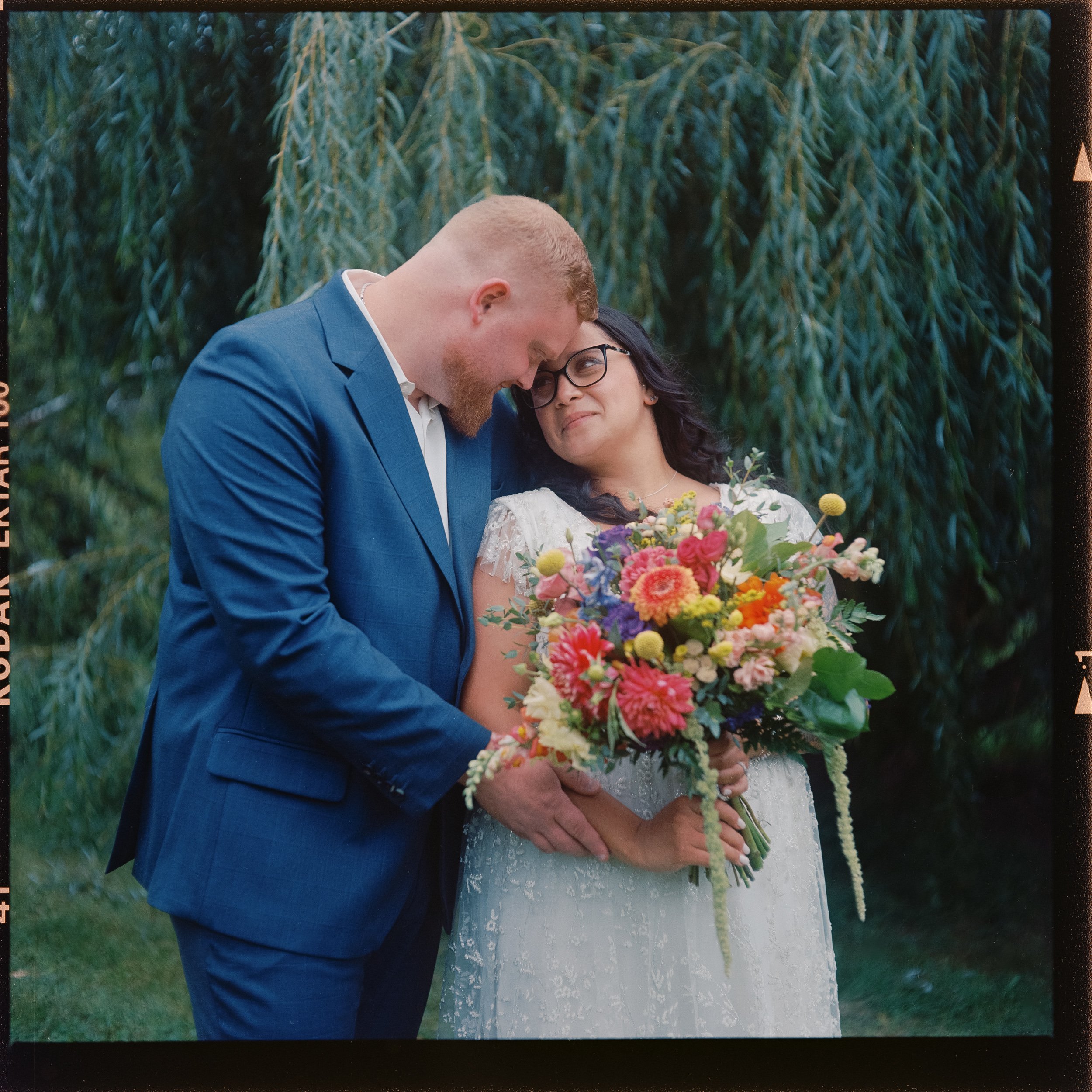 A bride and groom sharing an intimate moment outdoors, with the groom gently holding the bride's hands as she holds a colorful bouquet of flowers, against a backdrop of green trees.