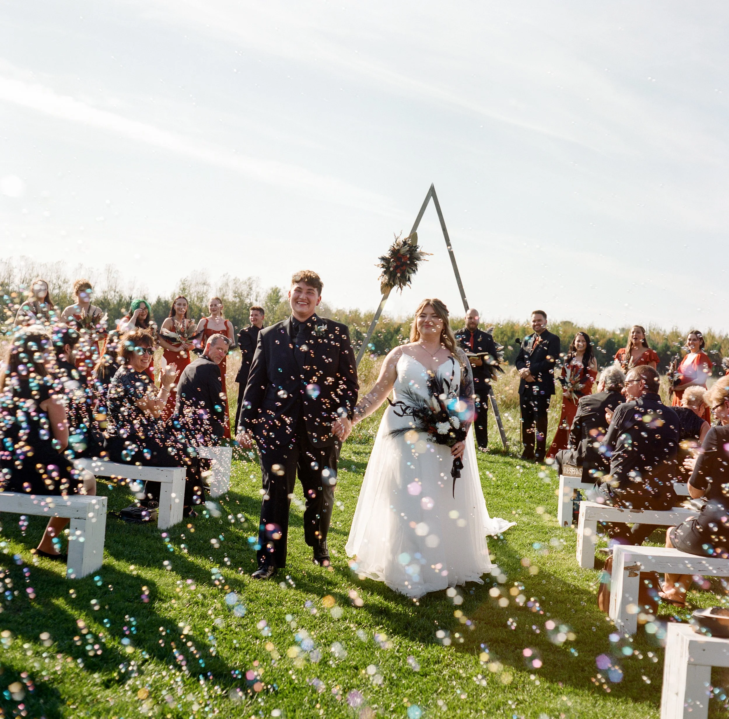 A wedding ceremony outdoors with a bride and groom holding hands while walking down the aisle amidst confetti, with guests seated on white benches on grassy land and an arch decorated with flowers in the background.