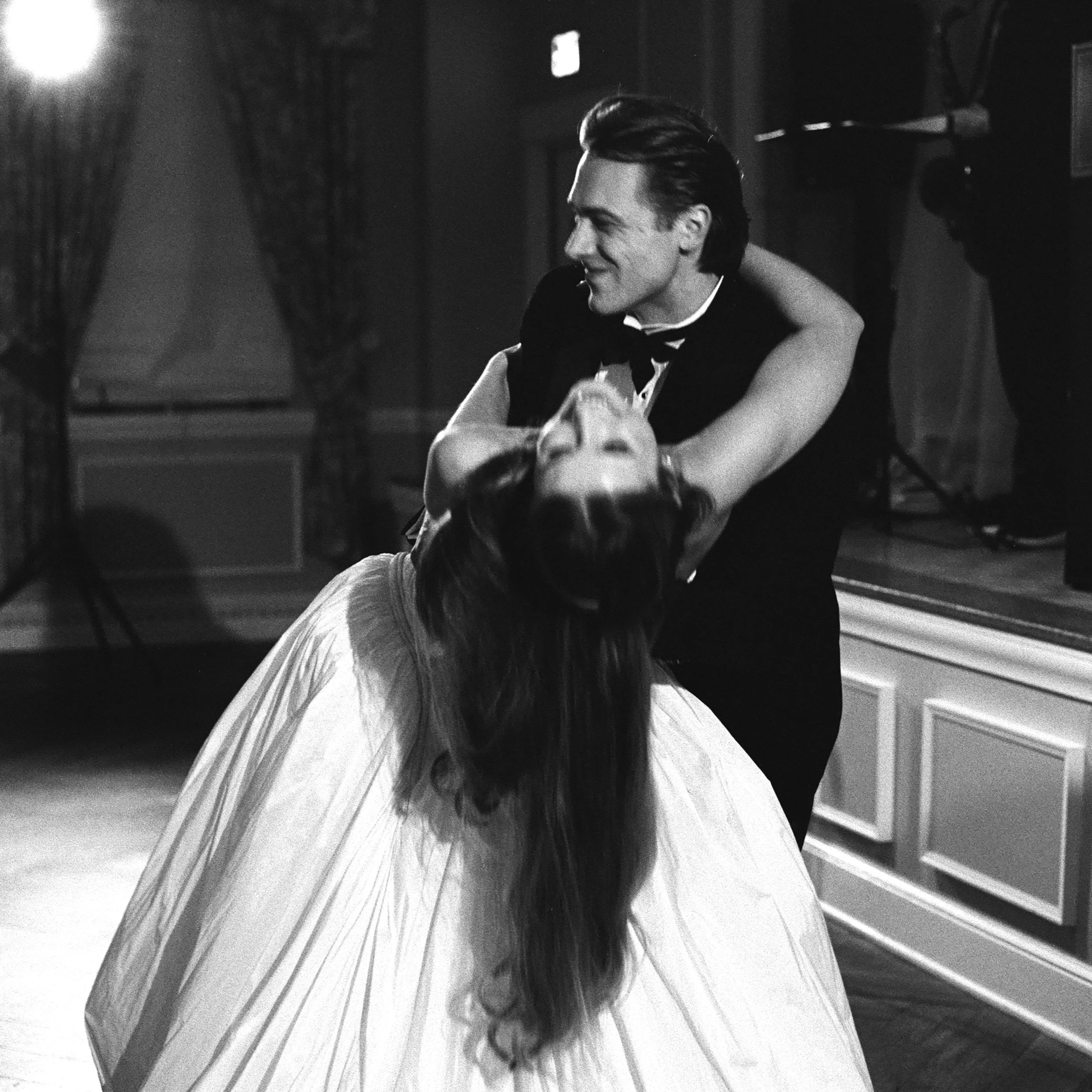 A man in tuxedo dancing with a woman in a white dress, holding her in a dance pose in a dimly lit room.
